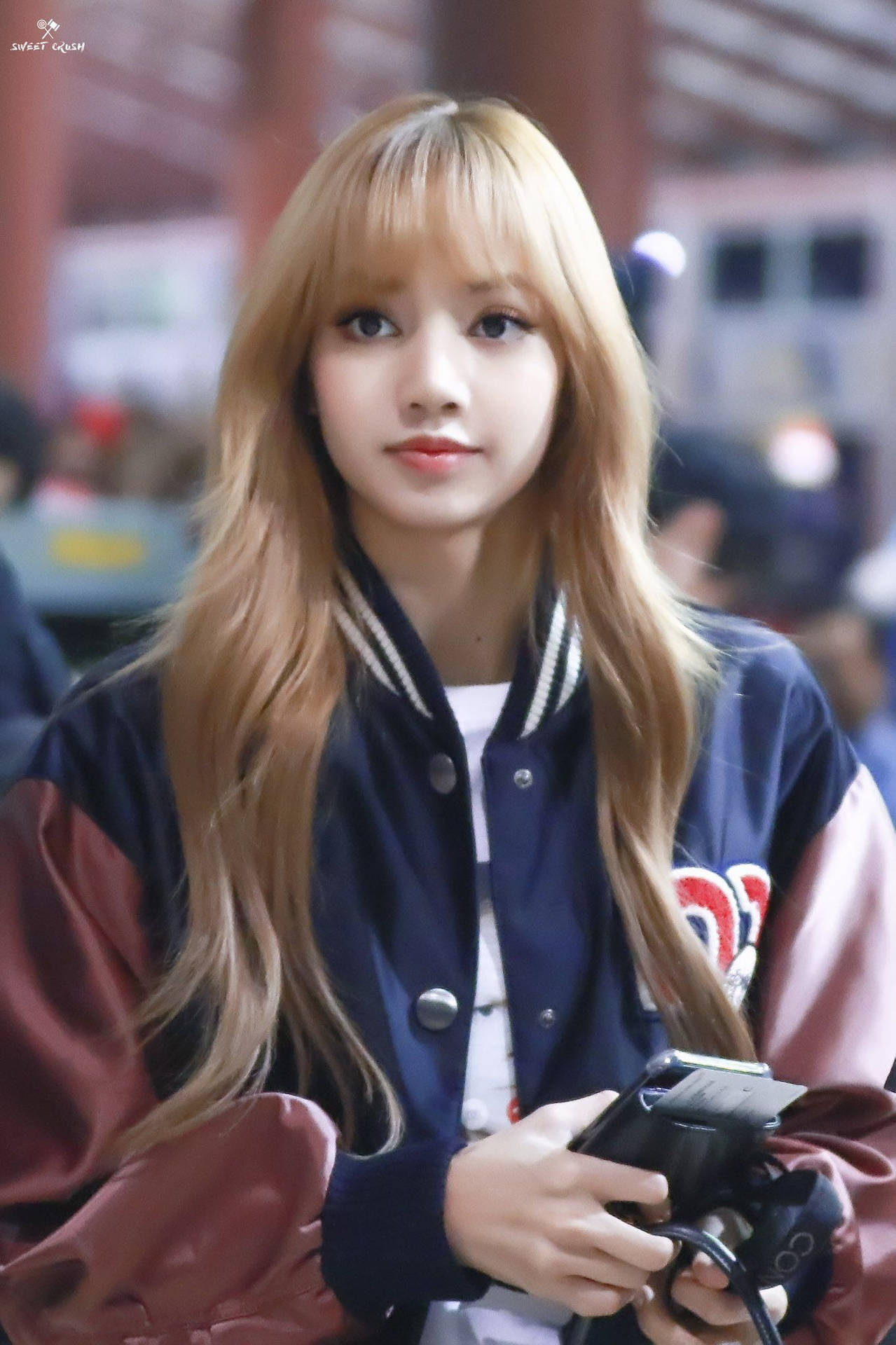 金髮年輕女性穿著校隊夾克在室內拿著手機。 — BLACKPINK Lisa GIF 壁紙