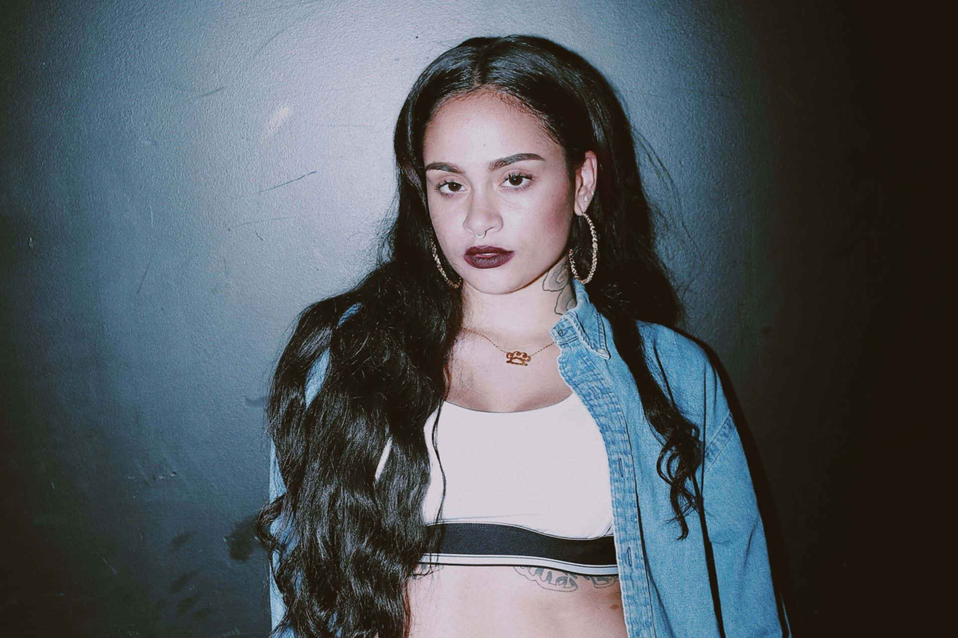 kehlani 桌布 壁紙 — 一位年輕女性，長長的黑髮和鮮明的妝容，在昏暗的灰色牆壁前直視鏡頭。