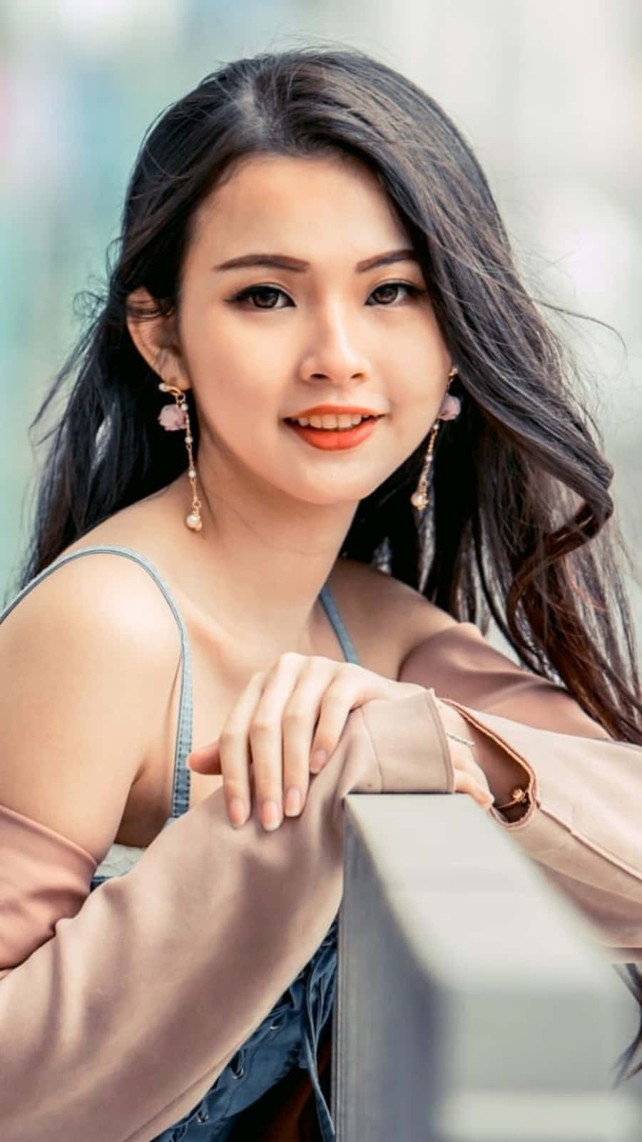 美女 桌布 壁紙 — 一位年輕女性，長長的黑髮和鮮豔的橙色唇膏，在柔和日光中微笑著靠在欄杆上。