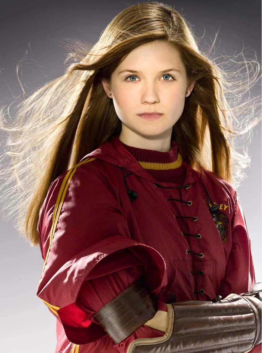 Ginny Weasley 桌布 壁紙 — 一位年輕女性，飄逸的金色長髮，身穿栗色魁地奇長袍和皮革護臂，背景為灰色攝影棚。