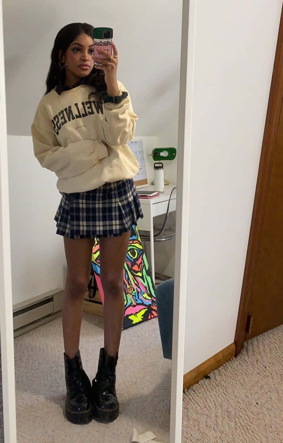 emo 美學 桌布 壁紙 — 一位年輕女性在休閒房間裡穿著奶油色衛衣、格子裙和厚底靴子對著鏡子自拍。