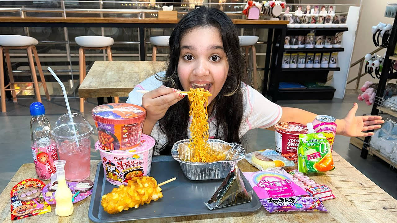 女孩 桌布 壁紙 — 一位年輕女性在休閒餐廳裡，熱情地吃著一大疊即食麵，周圍擺滿了色彩繽紛的韓國零食和飲料。