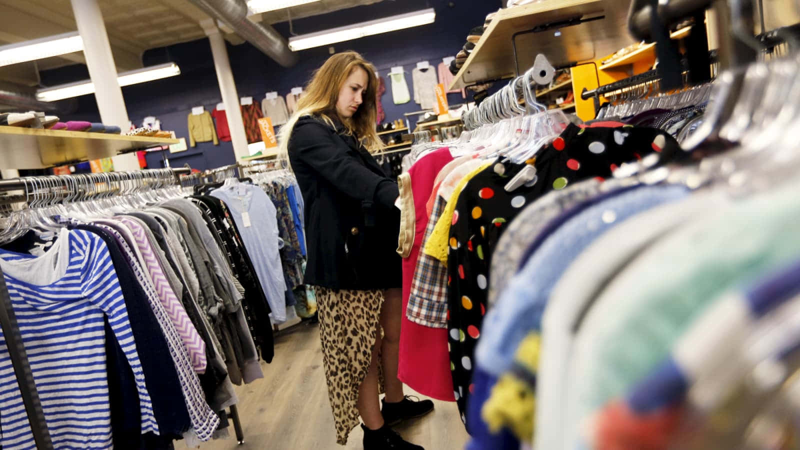 Shopping 桌布 壁紙 — 一位年輕女性在光線充足、有木地板和掛著衣服的二手店裡瀏覽色彩繽紛的衣架。