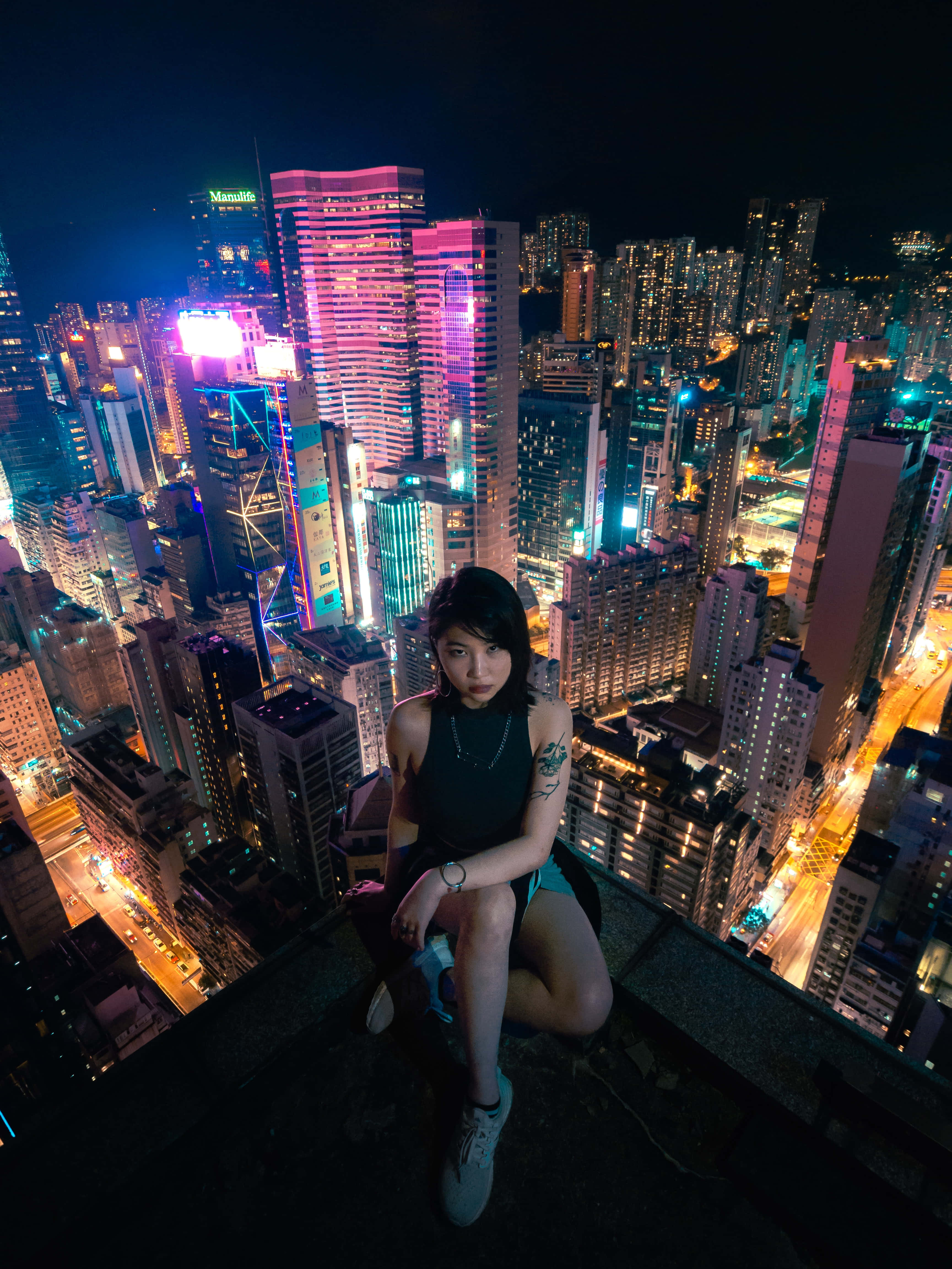 Rooftop 桌布 壁紙 — 一位年輕女性在夜晚坐在摩天大樓頂端，俯瞰著耀眼的霓虹燈城市景觀，高聳的建築物和發光的街道。