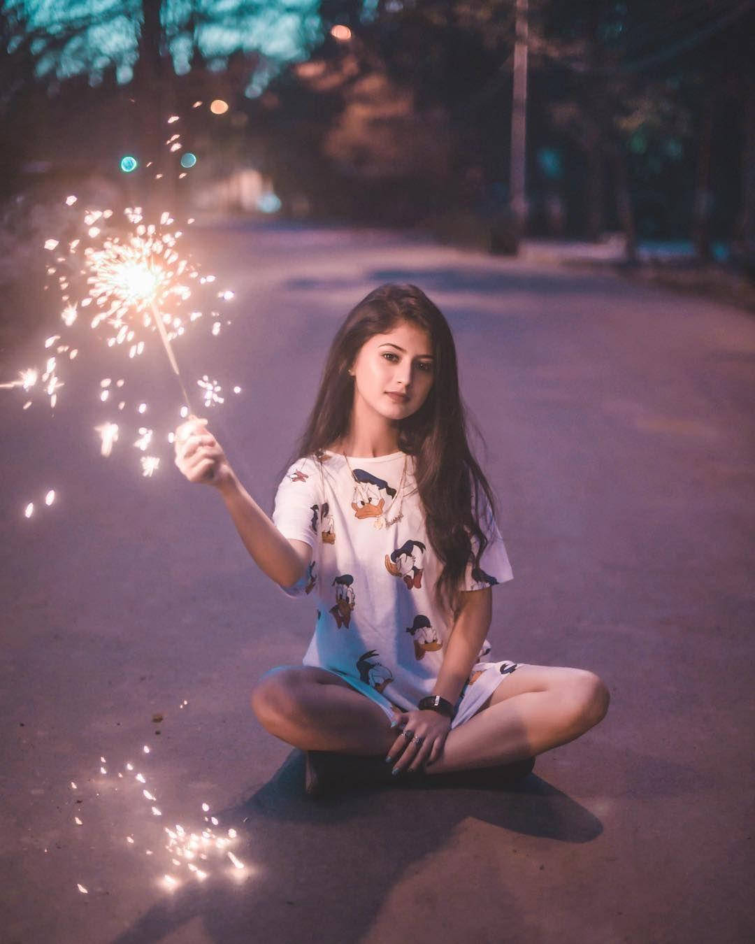 模特兒 桌布 壁紙 — 一位年輕女性在夜晚的街道上盤腿而坐，手持閃爍的煙火，溫暖的光芒照亮她的臉龐。