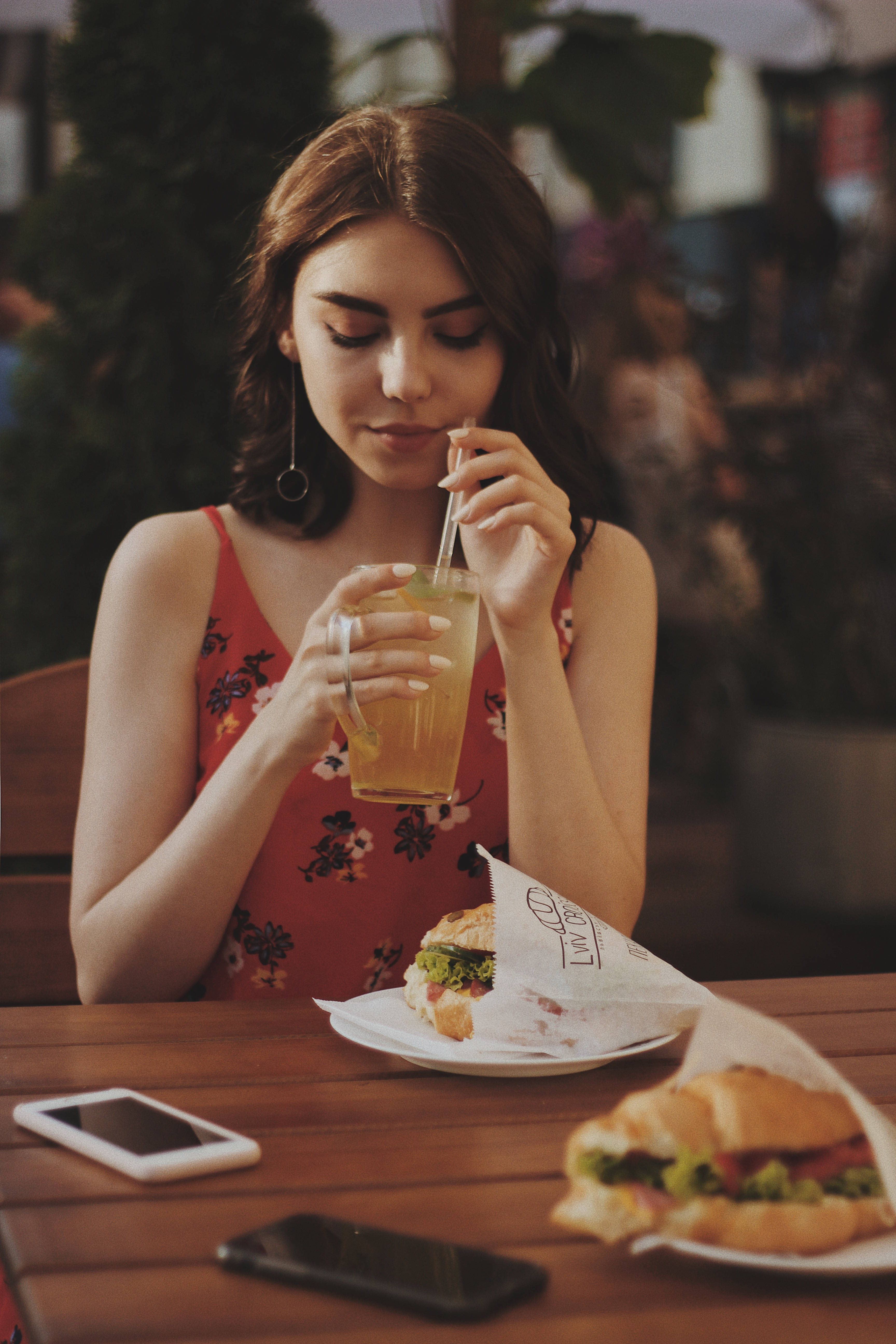 food iPhone 桌布 壁紙 — 一位年輕女性在戶外咖啡桌旁啜飲清涼飲品，旁邊放著三明治和電話。