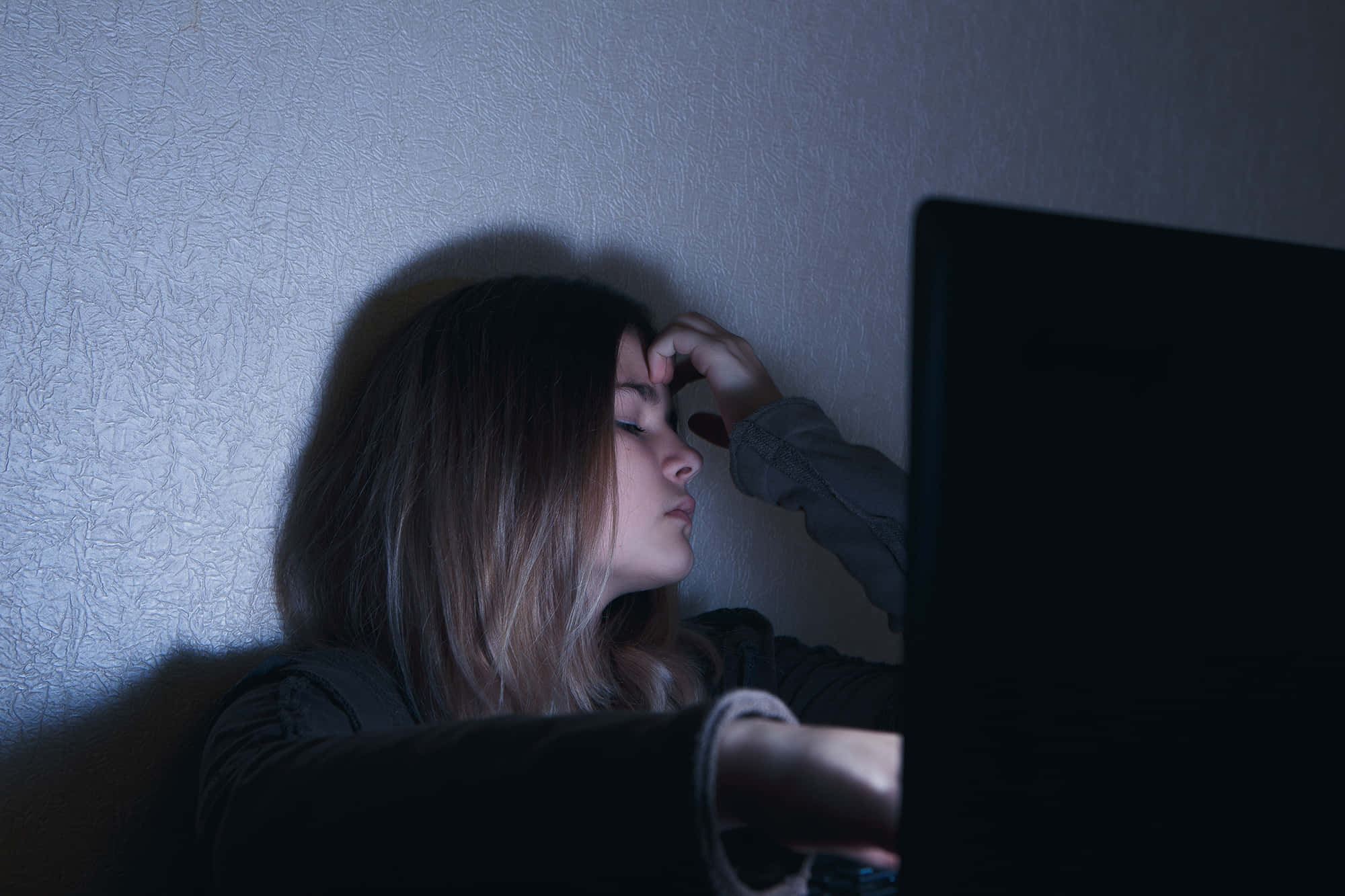 Depression Girl 桌布 壁紙 — 一位年輕女性在昏暗光線中靠著牆，手放在額頭上，因盯著筆記型電腦螢幕而疲憊不堪。