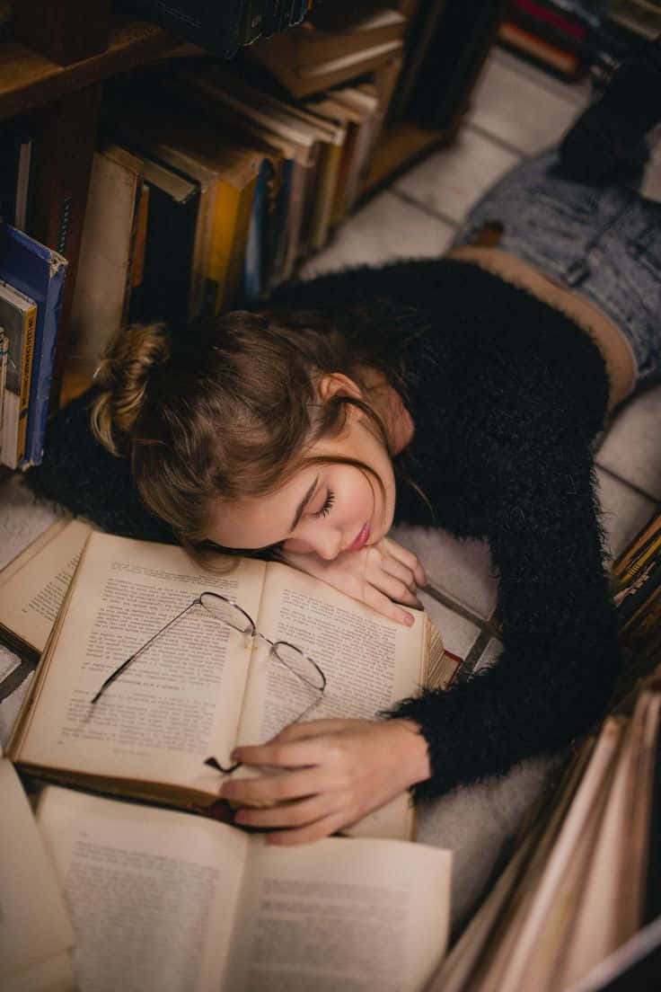 Aesthetic Cute Profile 桌布 壁紙 — 一位年輕女性在舒適的圖書館中，於高聳書架間的翻開書本上安詳入睡。