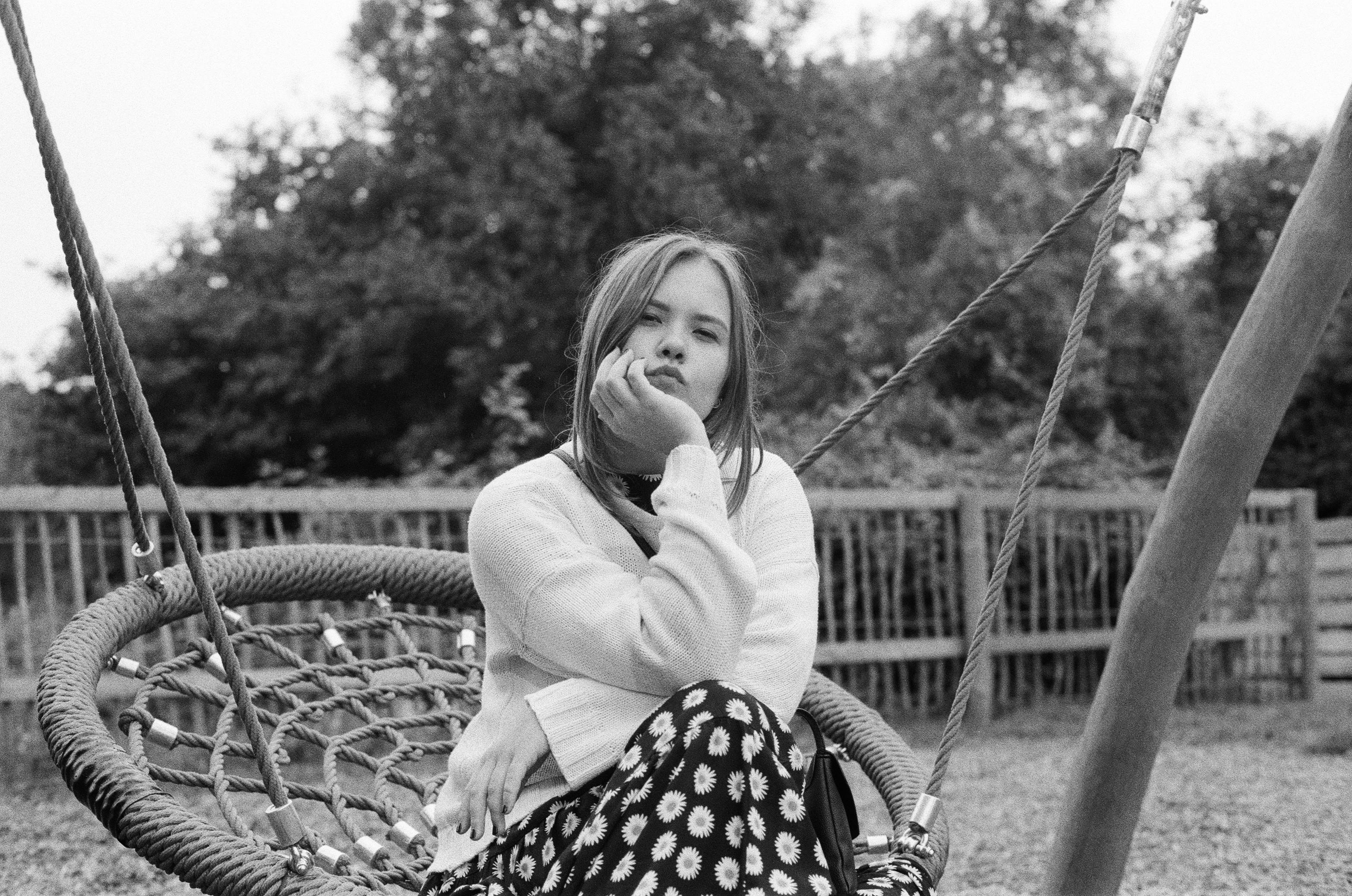 Playground 桌布 壁紙 — 一位年輕女性坐在戶外的繩索鞦韆上，手托下巴沉思，背景是樹木和木柵欄，以單色呈現。