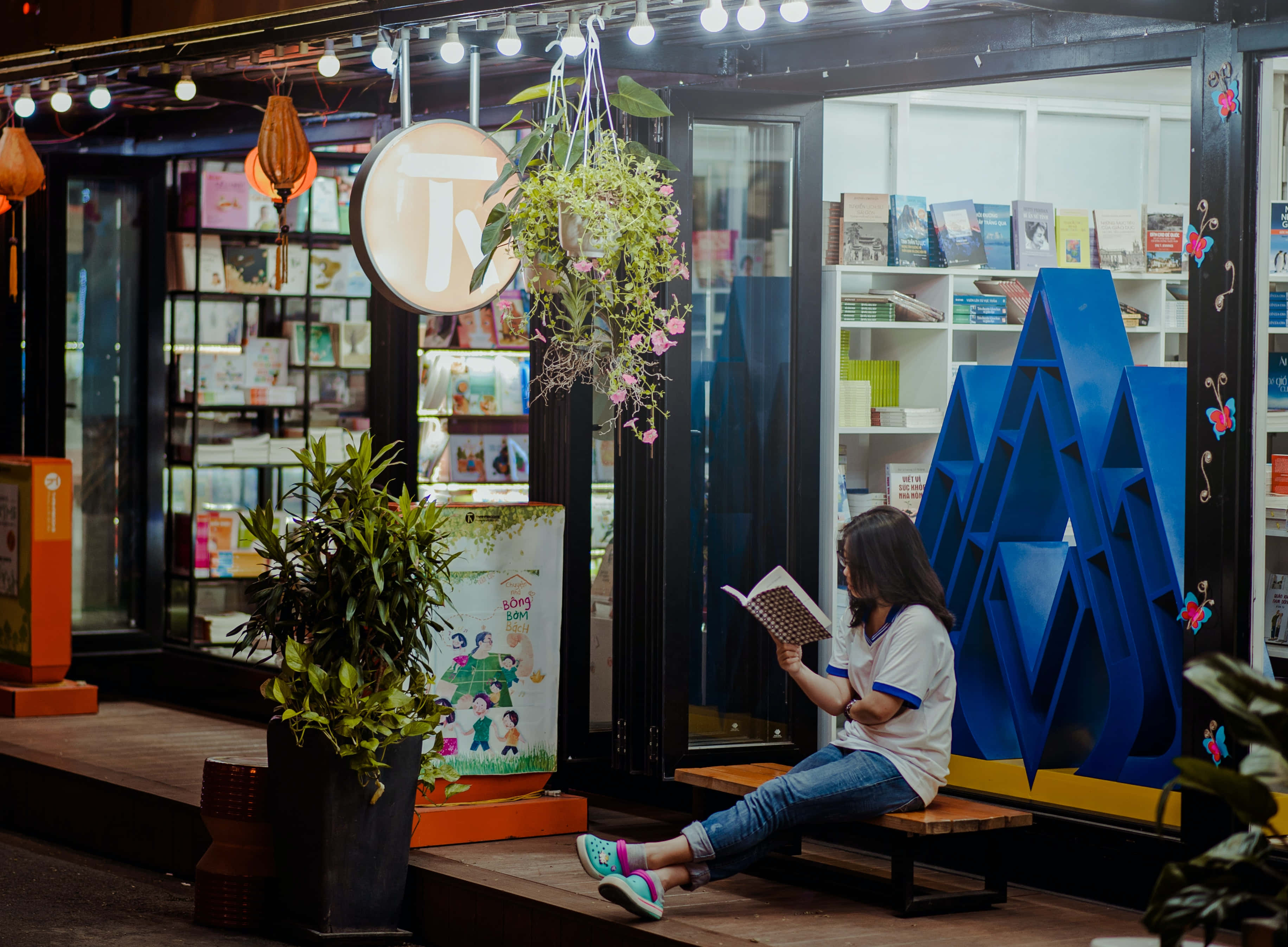 Store 桌布 壁紙 — 一位年輕女性坐在舒適書店外的木長椅上，在溫暖的串燈下閱讀一本書，周圍有懸掛植物和色彩繽紛的裝飾。