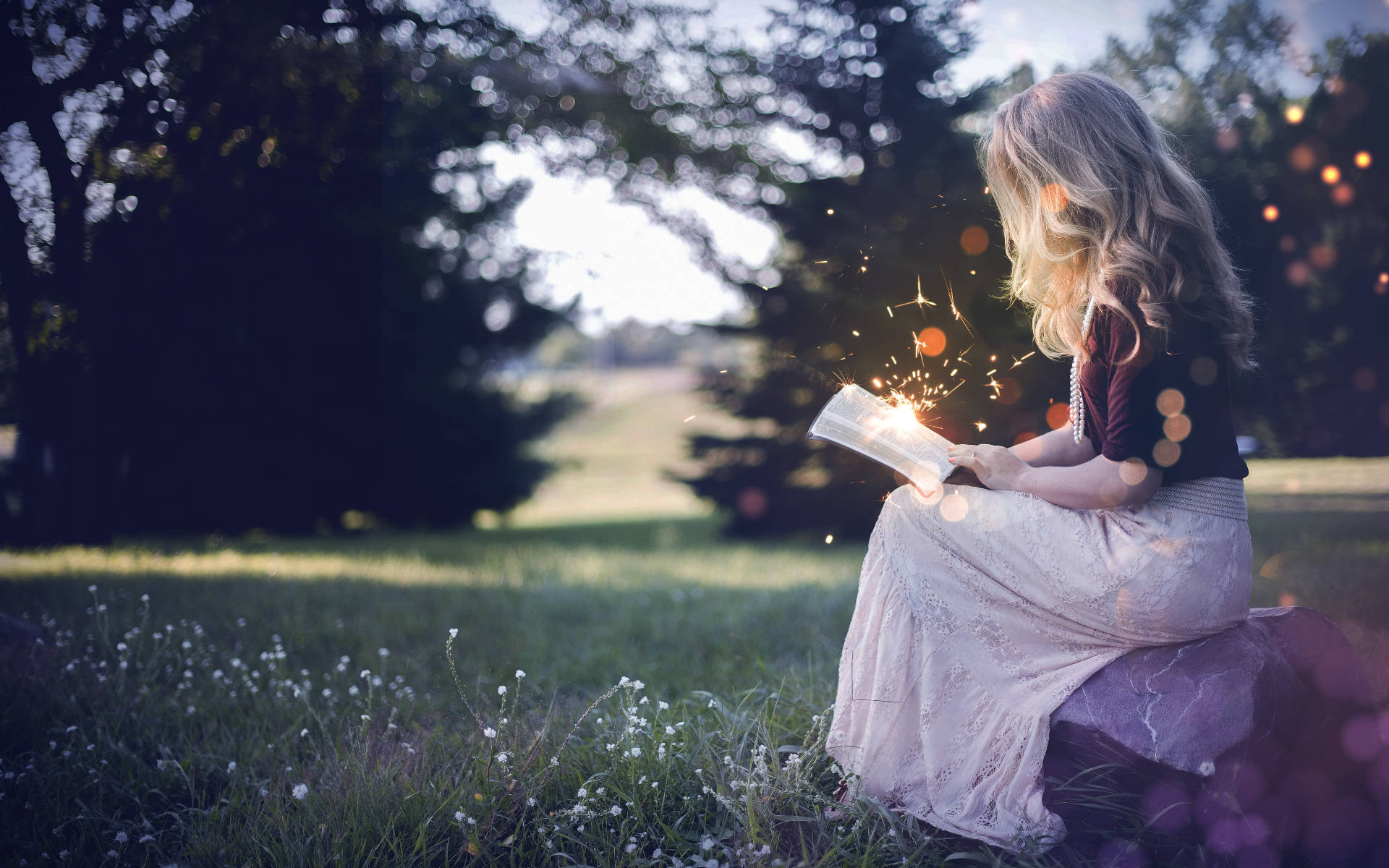 Reading 桌布 壁紙 — 一位年輕女性坐在陽光明媚的草地上，閱讀一本散發魔法火花和發光球體的書。