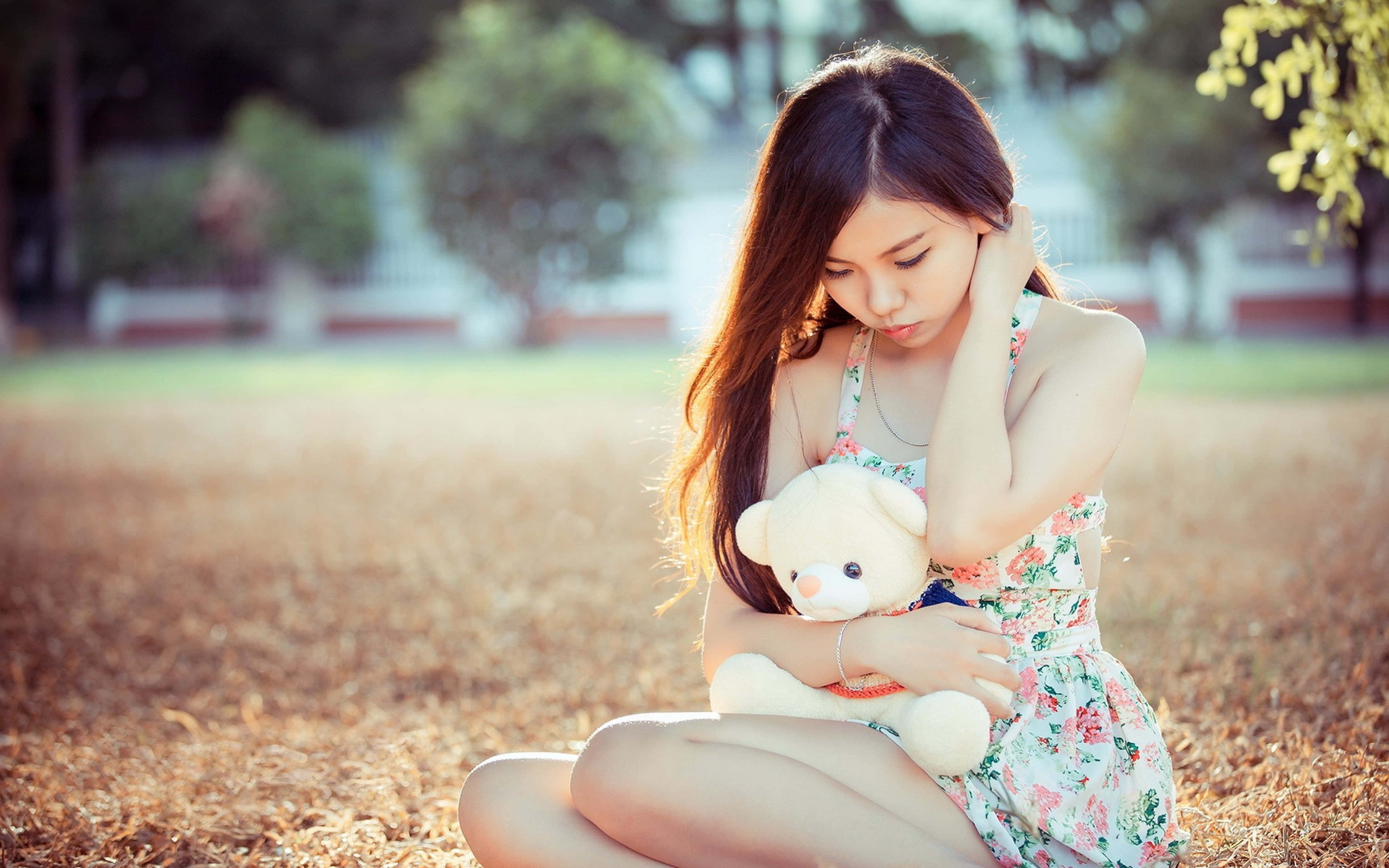 Teddy Bear 桌布 壁紙 — 一位年輕女性坐在陽光普照的田野中，抱著一隻穿著花卉連衣裙的泰迪熊，散發柔和溫暖的光芒。