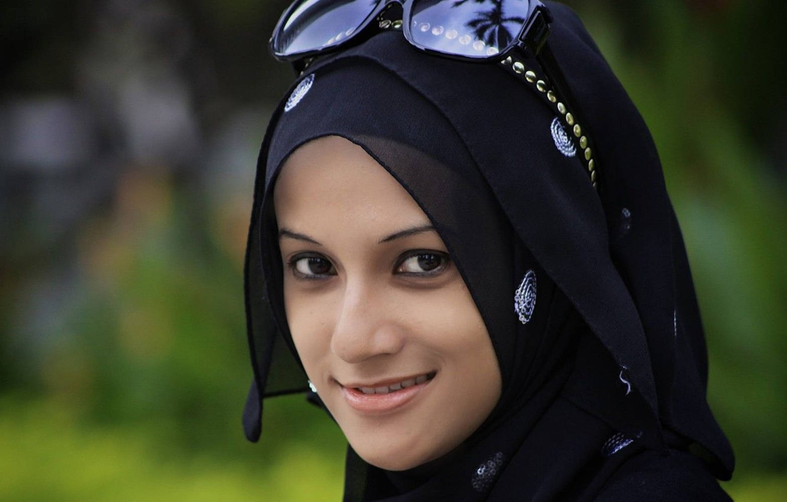 muslim 桌布 壁紙 — 一位年輕女性戴著帶有銀色刺繡的黑色頭巾，太陽眼鏡擱在頭上，在柔和的綠色模糊背景前溫柔微笑。