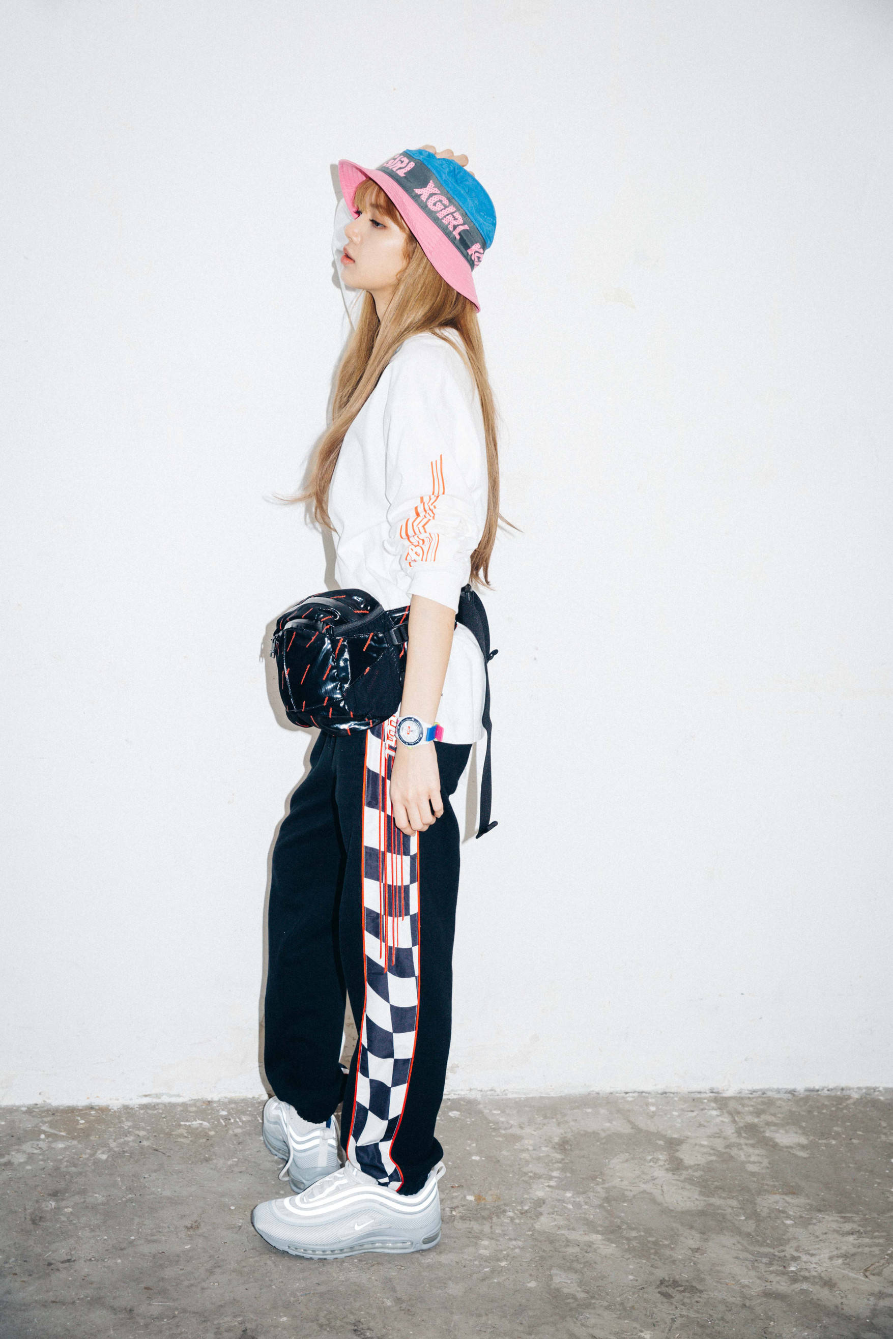 Lalisa 桌布 壁紙 — 一位年輕女性戴著彩色漁夫帽和格子褲，背著時尚包包和運動鞋，在純白牆前擺姿勢。