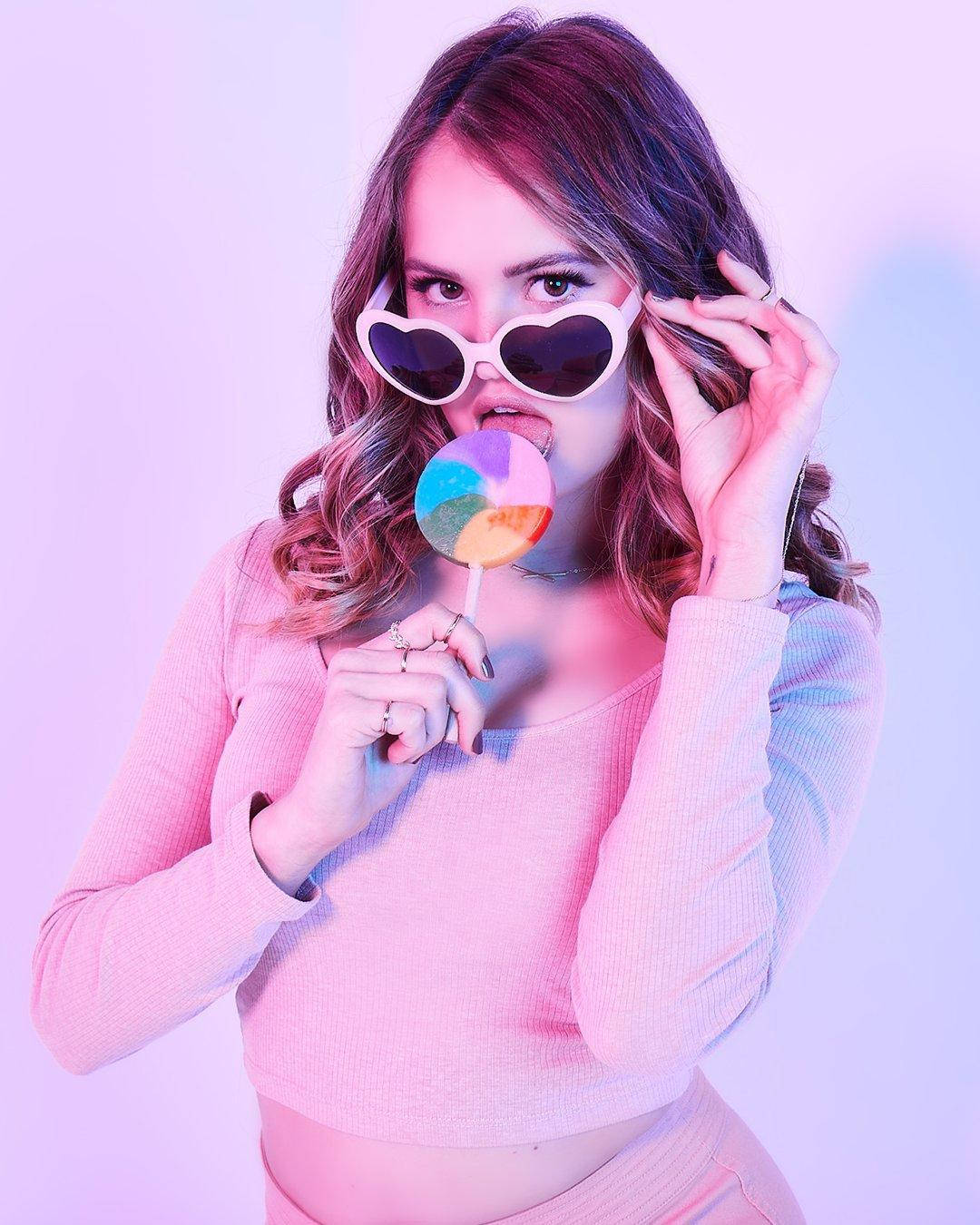 debby ryan 桌布 壁紙 — 一位年輕女性戴著心形太陽眼鏡，在柔和的粉紅和紫色漸變背景前咬著彩虹棒棒糖。