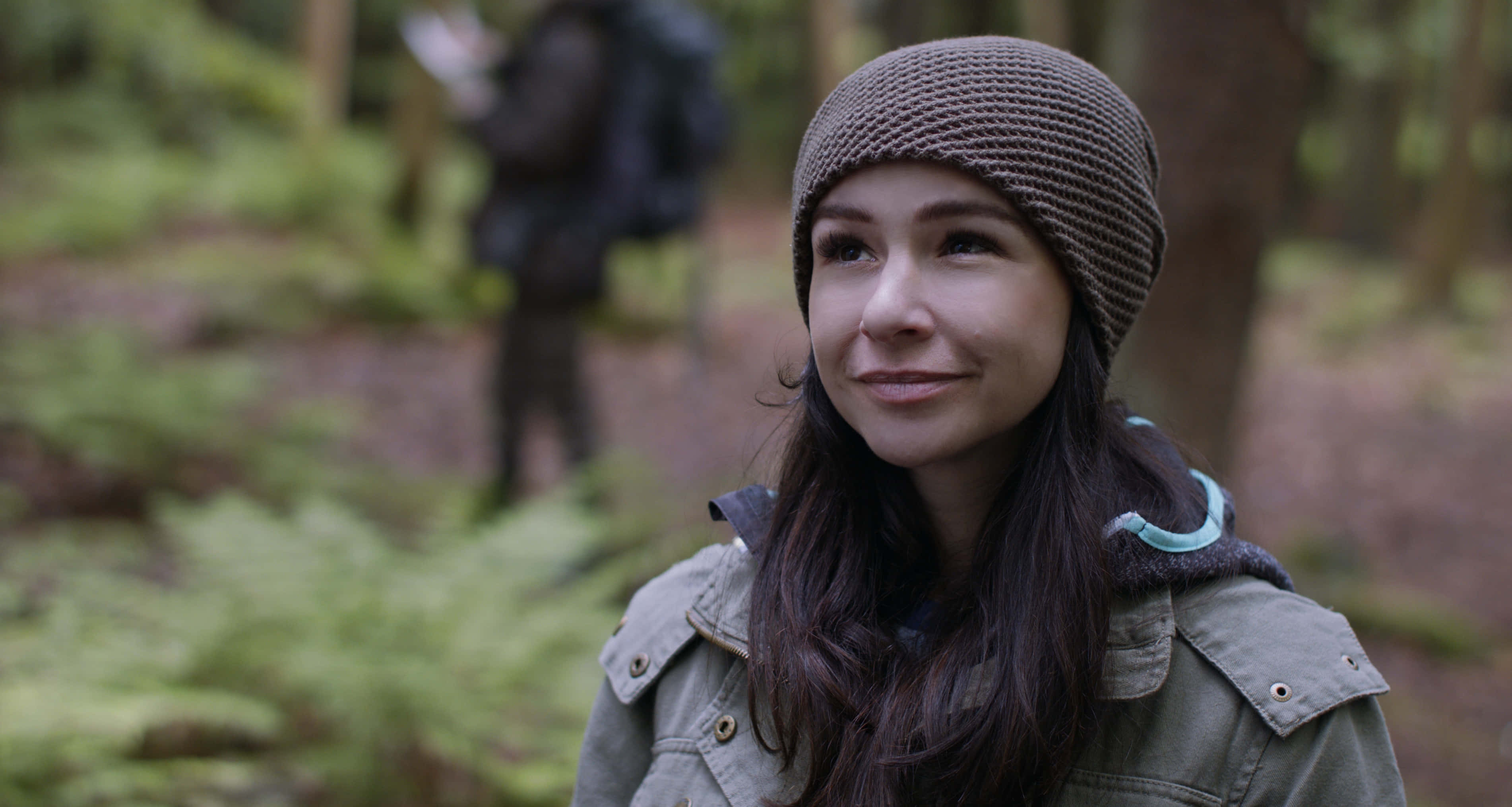 Danielle Harris 桌布 壁紙 — 一位年輕女性戴著棕色針織帽和橄欖綠外套，在長滿蕨類植物和樹木的茂密綠色森林中微笑。