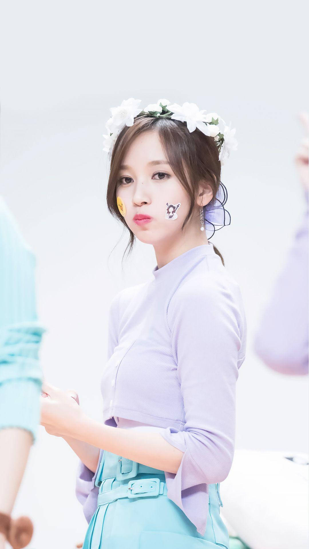 Kpop Aesthetic 桌布 壁紙 — 一位年輕女性戴著白色花冠和俏皮的面部貼紙，穿著柔和的薰衣草上衣和青綠色裙子，在明亮的白色背景前擺姿勢。