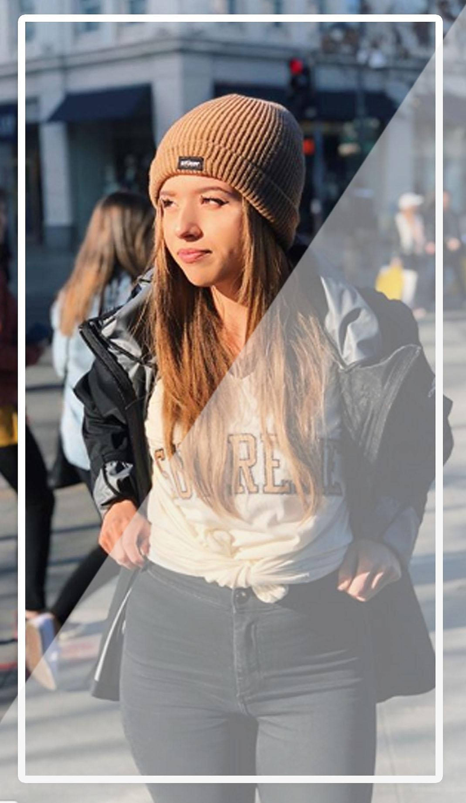 pokimane 桌布 壁紙 — 一位年輕女性戴著米色冷帽和黑色外套，自信地走在陽光普照的城市街道上，身後有模糊的行人。
