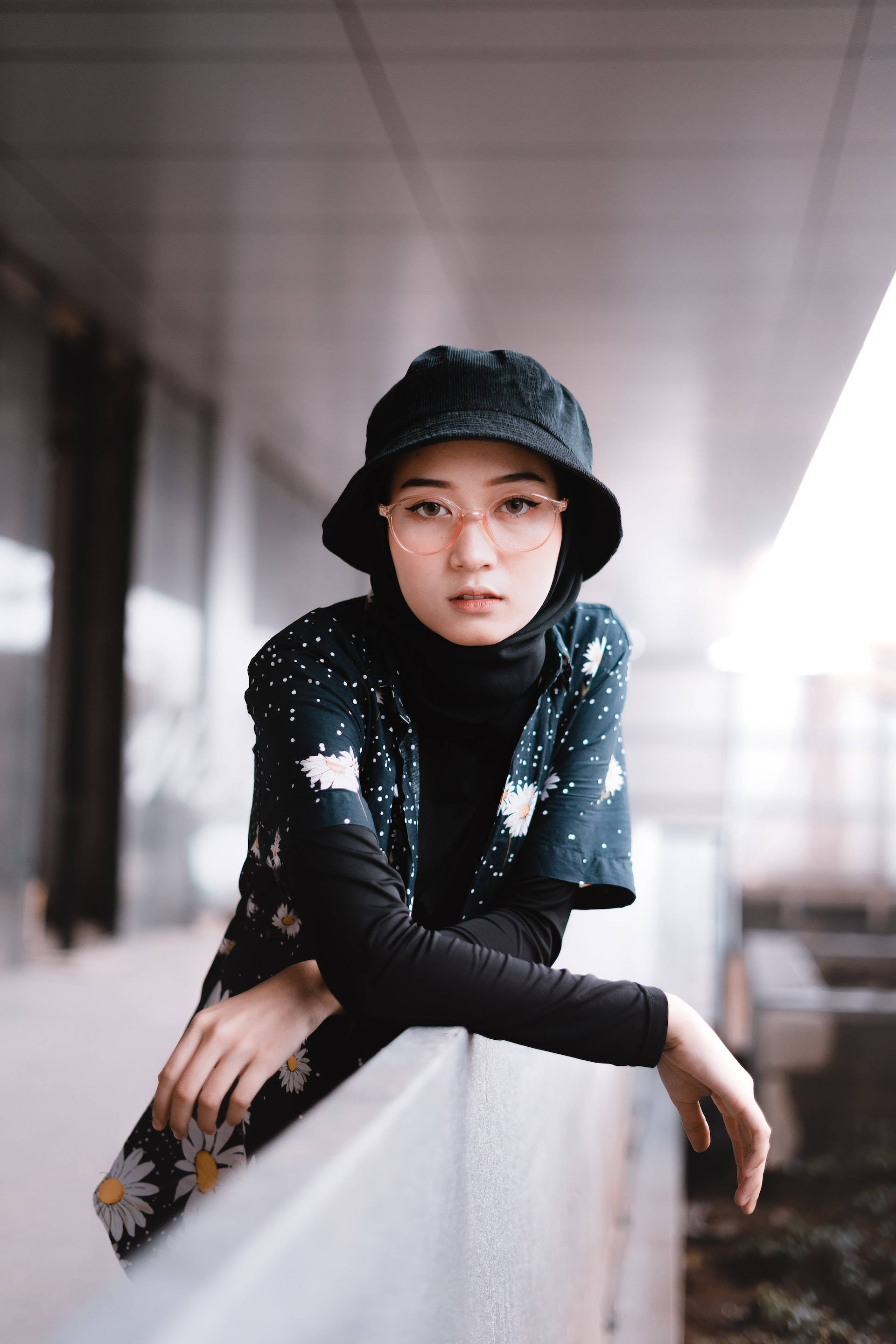 Asian Woman 桌布 壁紙 — 一位年輕女性戴著黑色漁夫帽和花卉頭巾，在柔和自然光下以嚴肅的神情倚靠在欄杆上。