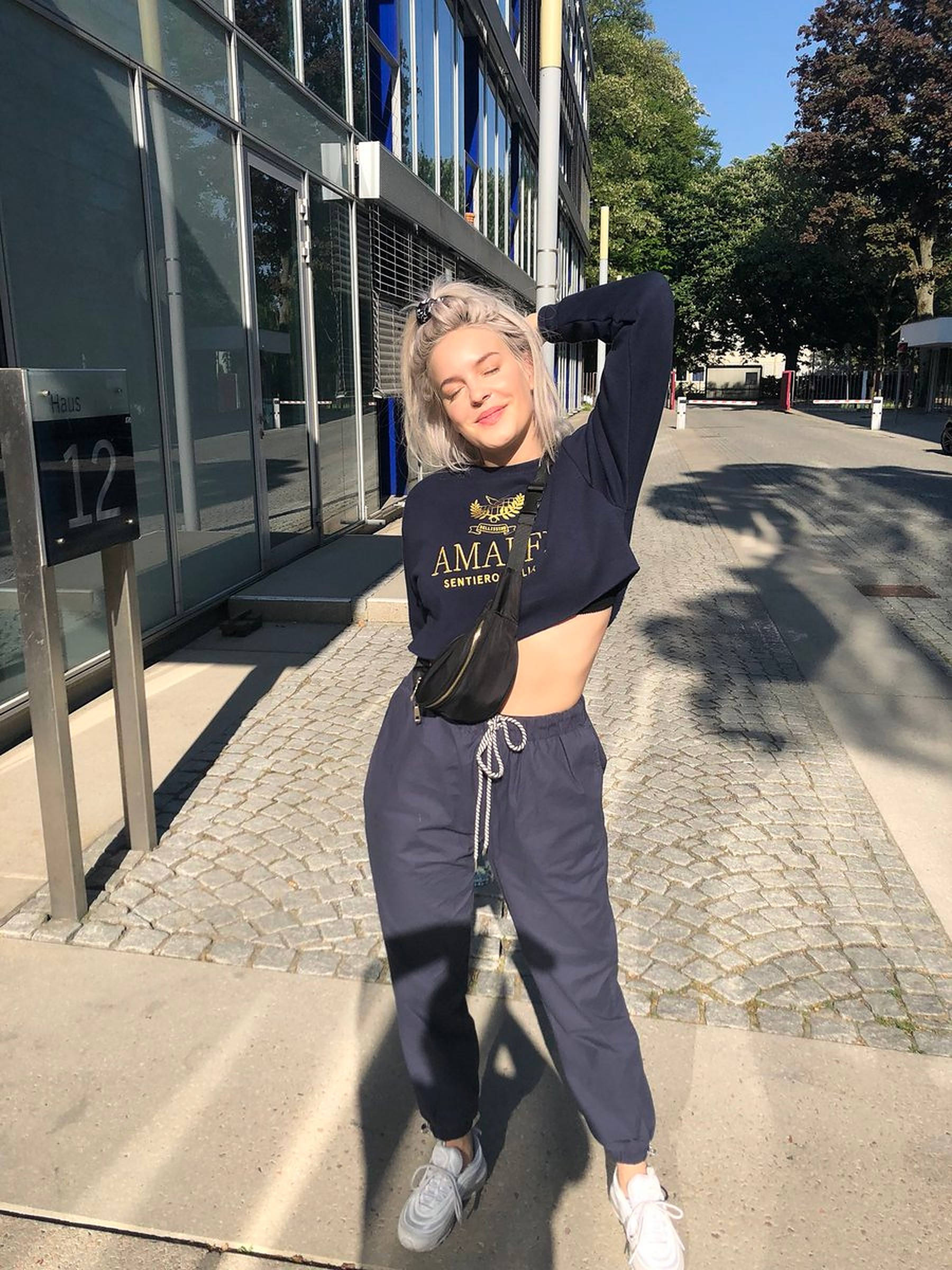 Anne Marie 桌布 壁紙 — 一位年輕女性擁有一頭鉑金色頭髮，在陽光明媚的城市人行道上自信地擺姿勢，穿著露臍上衣和運動褲。
