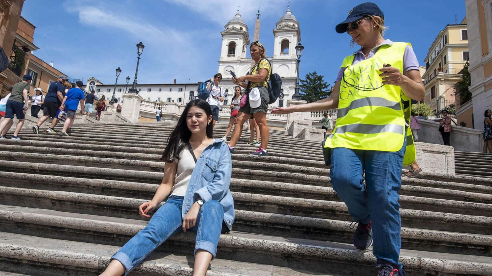 stairs 桌布 壁紙 — 一位年輕女性放鬆地坐在古老的石階上，附近一位穿著亮黃色背心的導遊在晴朗的藍天下做手勢。
