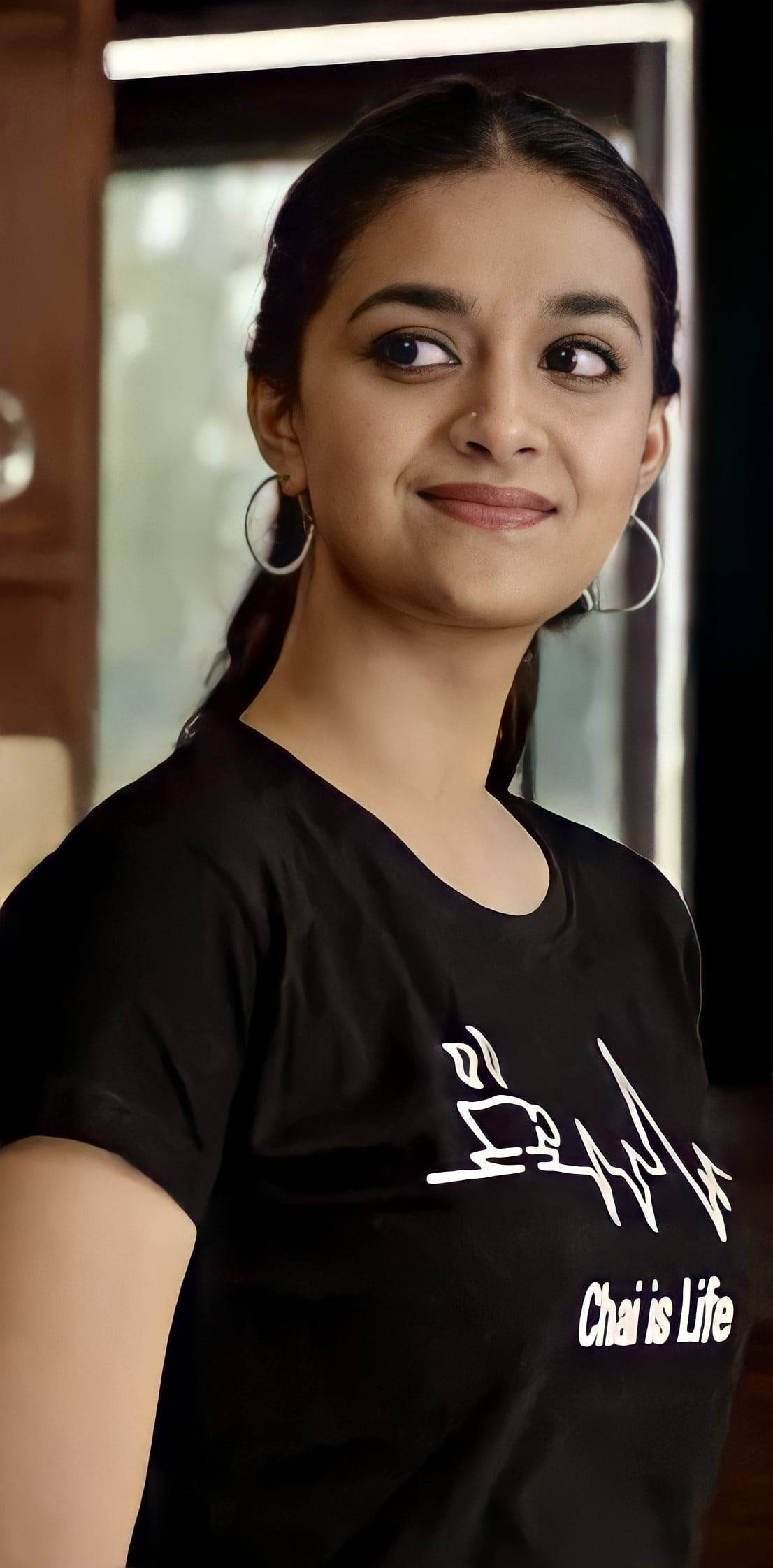 telugu 桌布 壁紙 — 一位年輕女性梳著辮子、戴著圓環耳環，穿著印有白色茶主題圖案和文字的黑色T恤，溫柔地微笑。