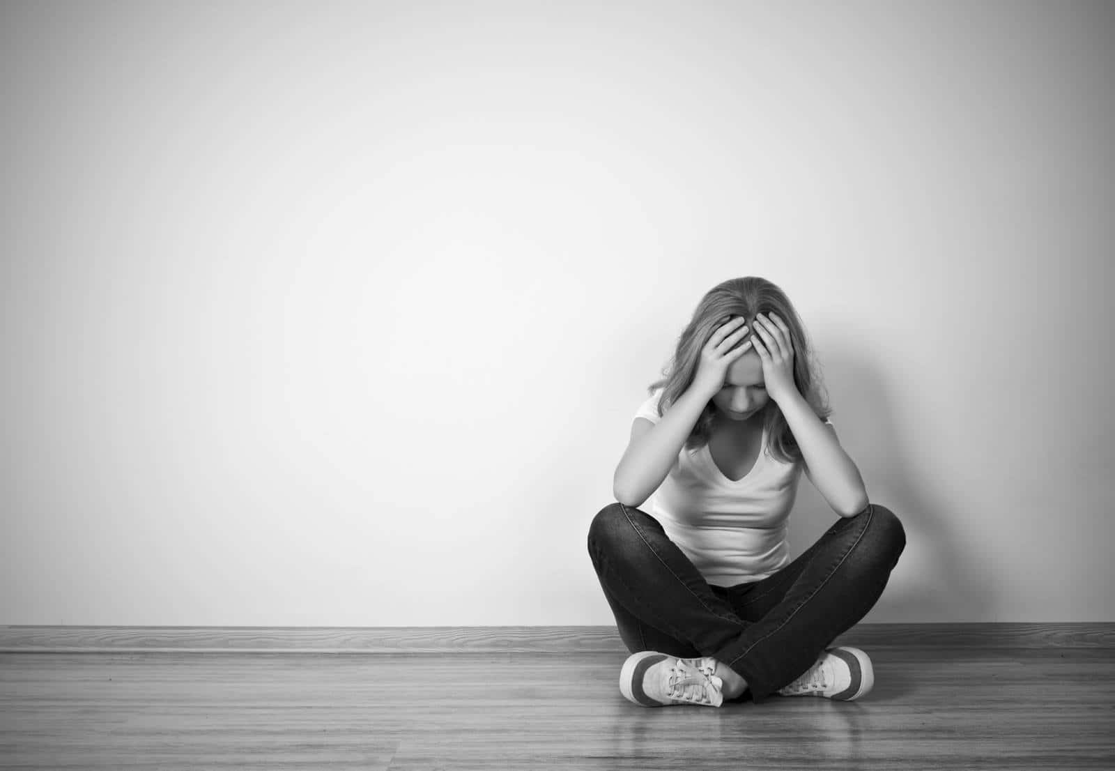 Depressed 桌布 壁紙 — 一位年輕女性獨自坐在木地板上，背靠素牆，雙手掩面，神情痛苦。