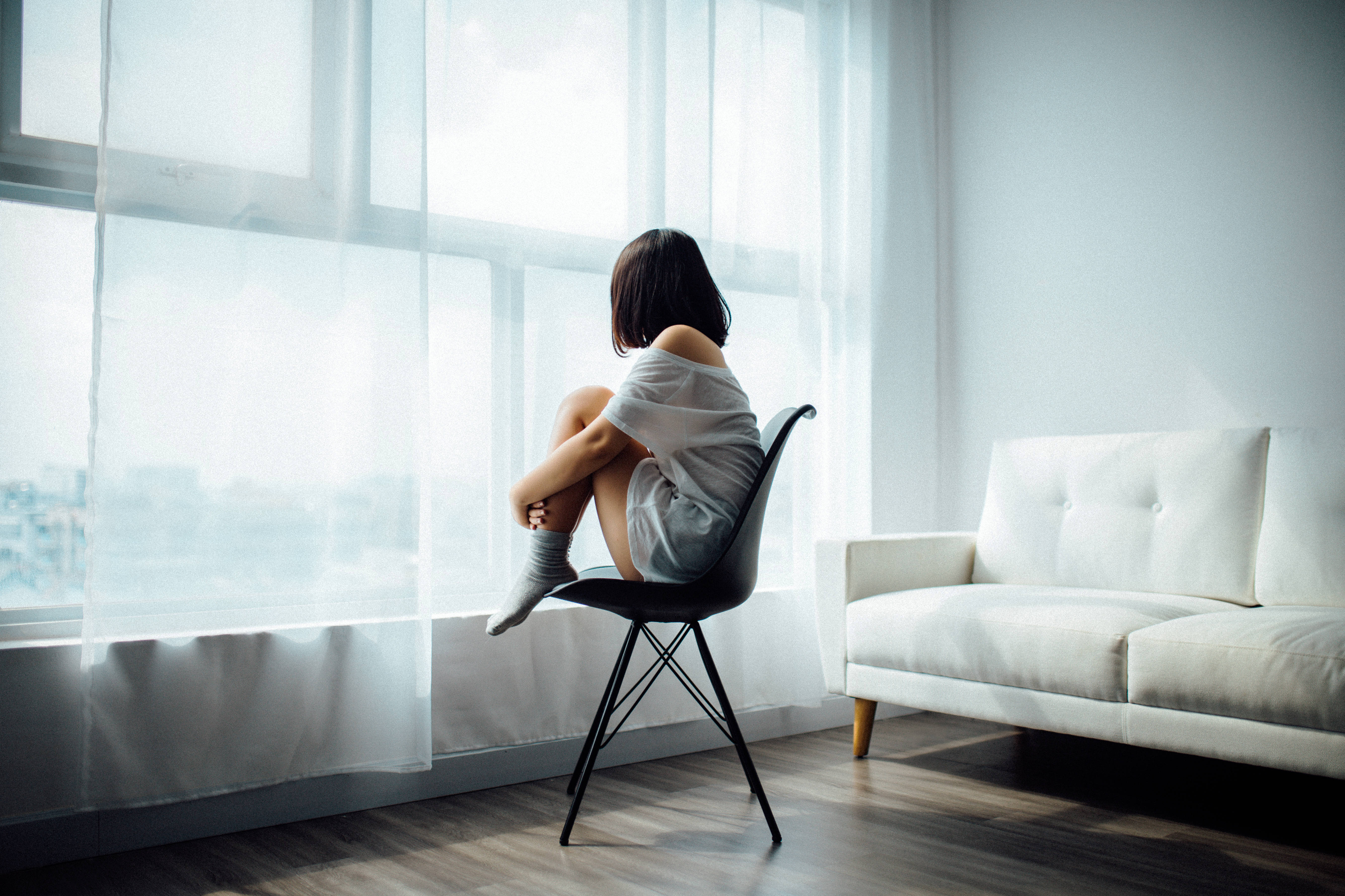 Alone 桌布 壁紙 — 一位年輕女性獨自坐在簡約白色客廳明亮窗邊的黑色椅子上，靜靜地凝視窗外。