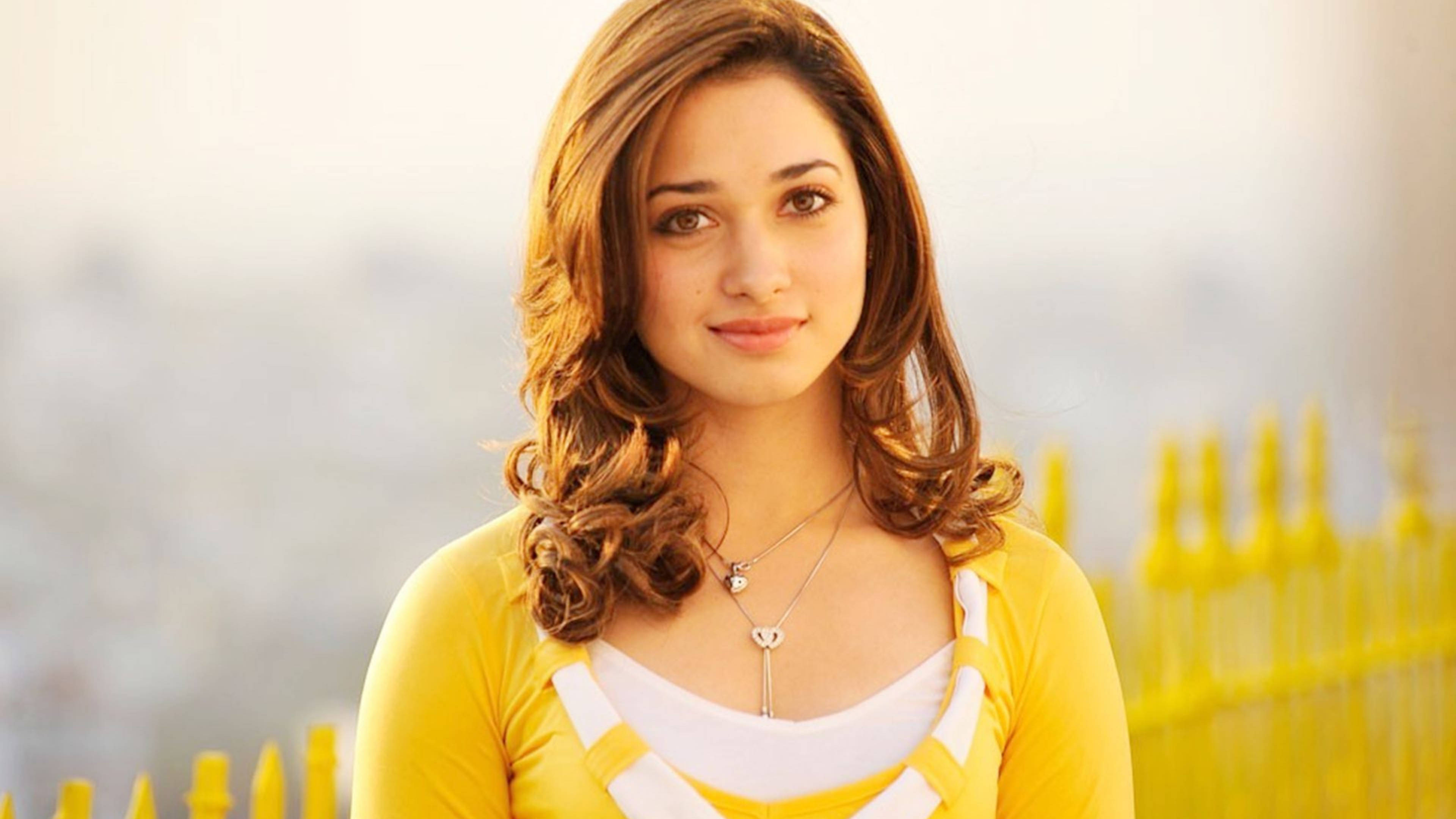tamannaah bhatia 桌布 壁紙 — 一位年輕女性留著波浪形棕色頭髮，穿著黃色開襟毛衣，在模糊的黃色籬笆背景下溫柔微笑。