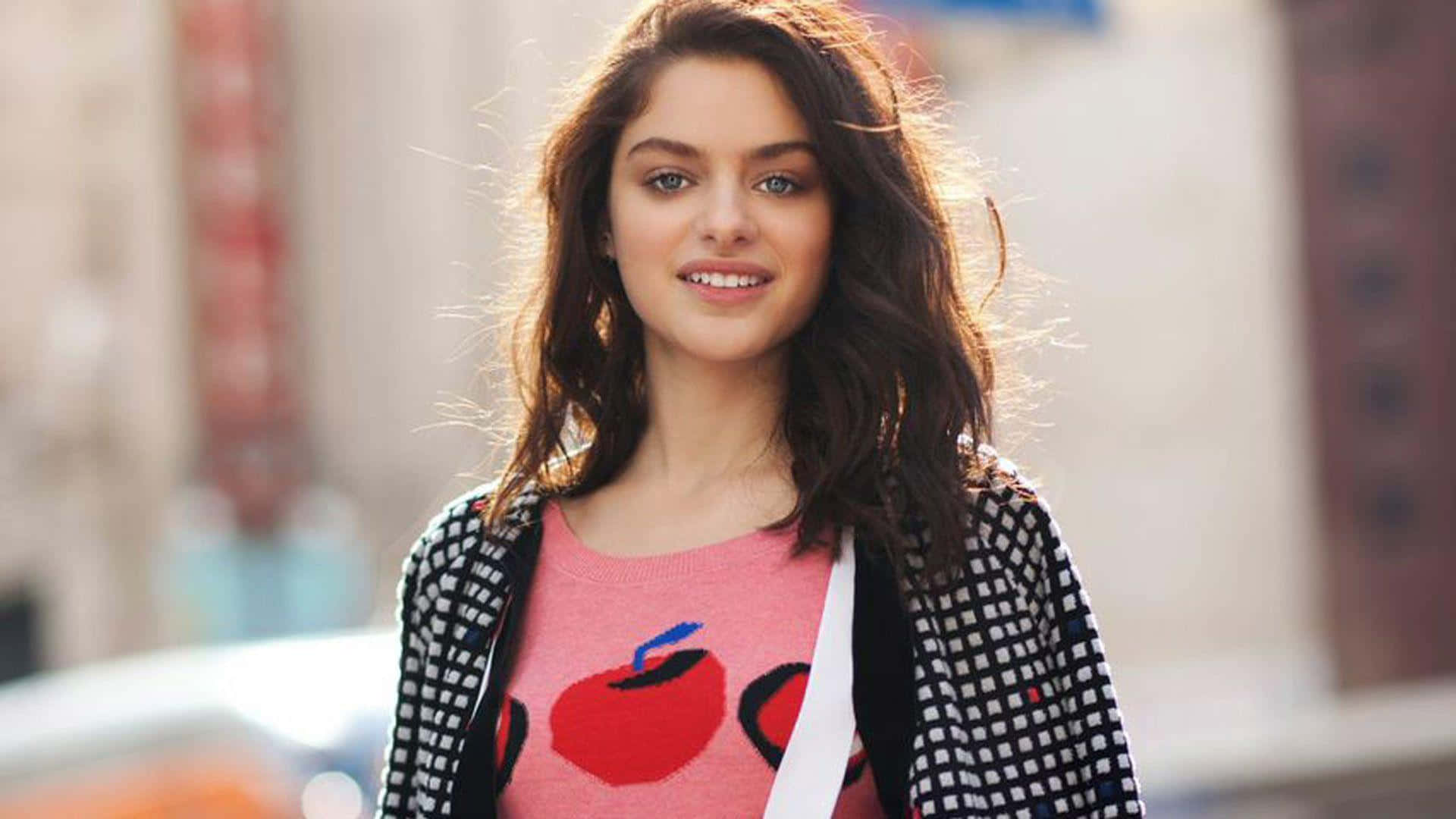 Odeya Rush 桌布 壁紙 — 一位年輕女性留著波浪棕色長髮，穿著粉紅色蘋果印花毛衣和格紋開襟衫，在城市的街道上溫暖微笑。