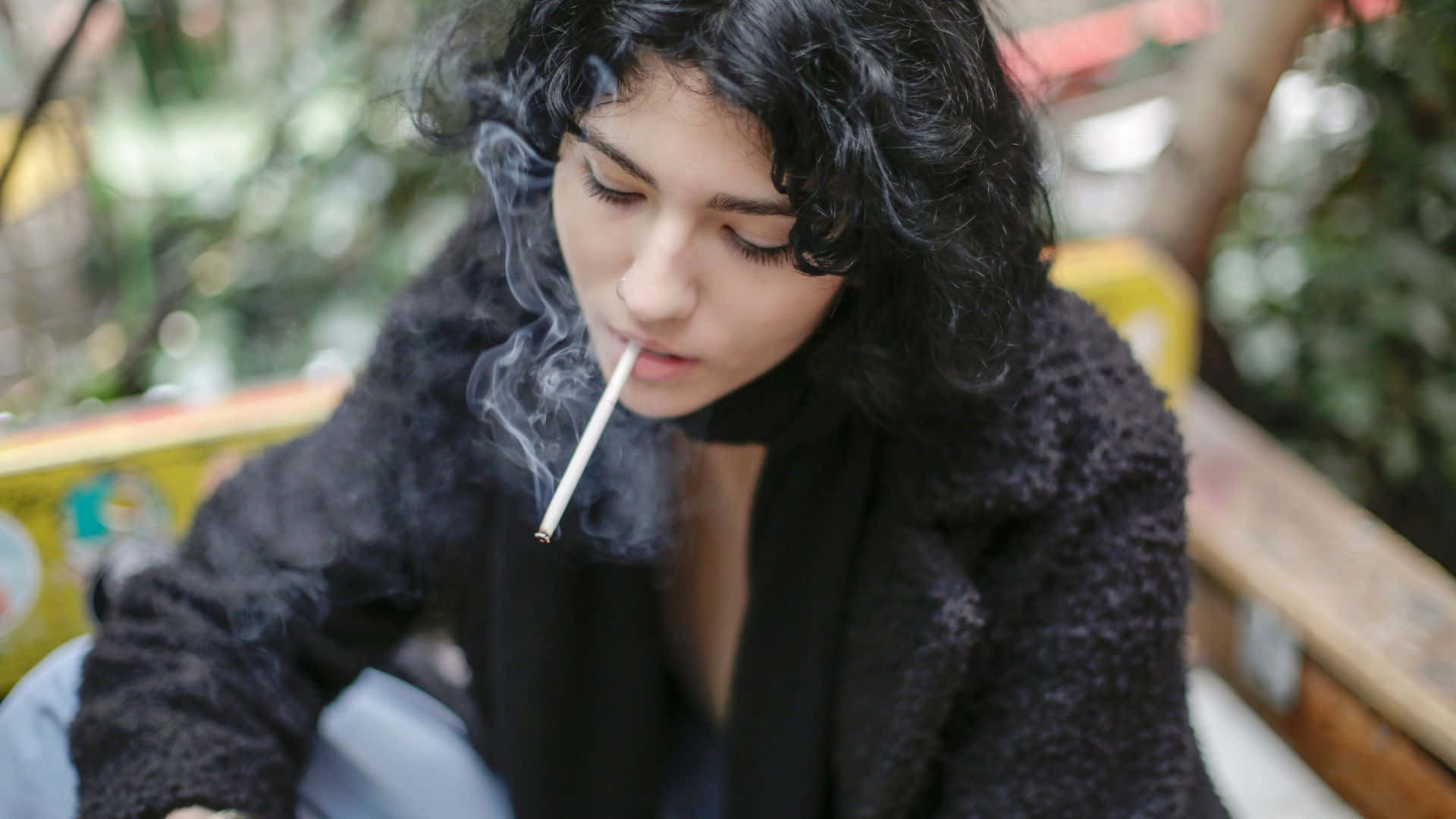 抽菸 桌布 壁紙 — 一位年輕女性留著深色捲髮，坐在戶外抽煙，煙霧在她臉龐縈繞。