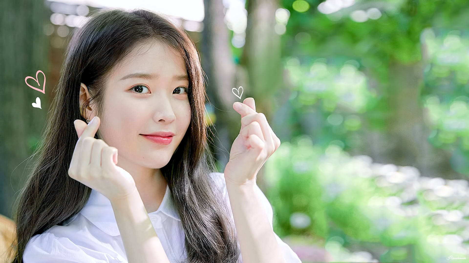 Kpop Aesthetic 桌布 壁紙 — 一位年輕女性留著深色長髮，在柔和的綠色森林背景下，雙手比出愛心手勢，溫柔微笑。