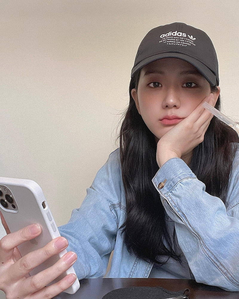 jisoo 可愛 桌布 壁紙 — 一位年輕女性留著深色長髮，戴著黑色 adidas 帽子和牛仔外套，一邊拿著手機，一邊用手托著下巴。