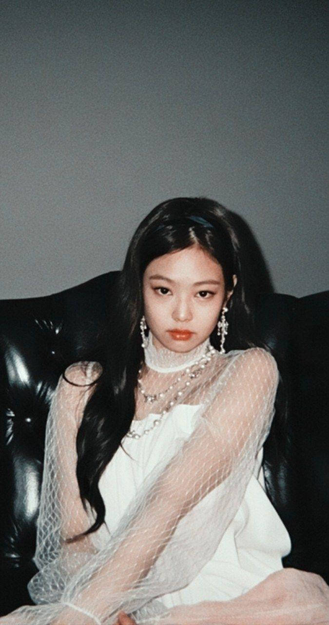 Jennie 桌布 壁紙 — 一位年輕女性留著深色長髮，端莊地坐在黑色皮革沙發上，身穿白色透視裝和珍珠首飾，背景是柔和的灰色牆壁。