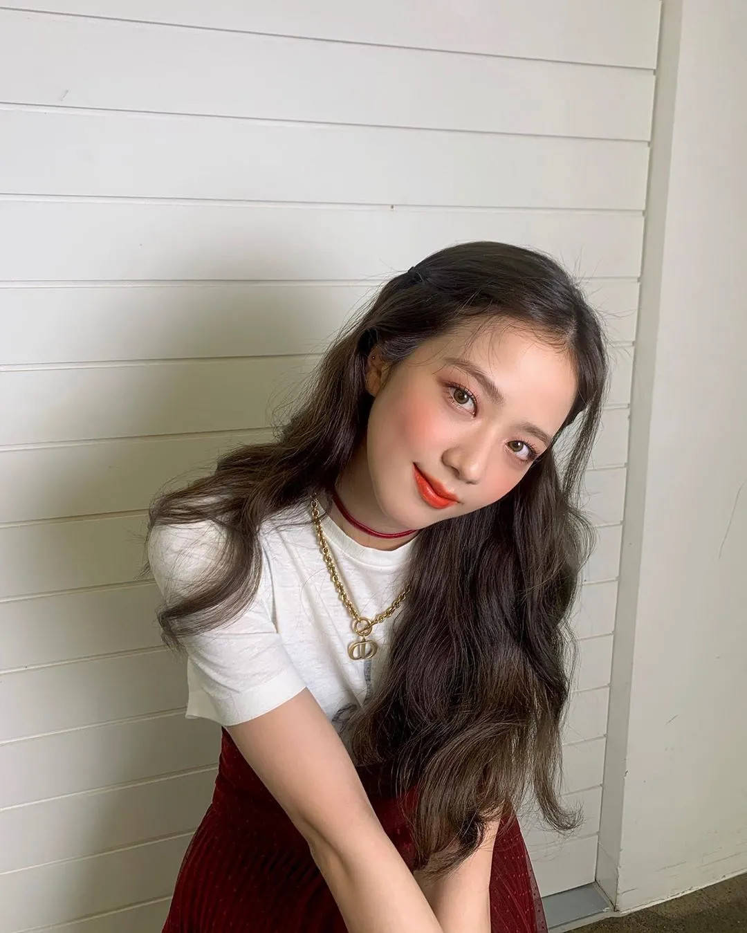 Jisoo Cute 桌布 壁紙 — 一位年輕女性留著深色長髮，身穿紅色連衣裙和金色項鍊，靠著白色木牆。