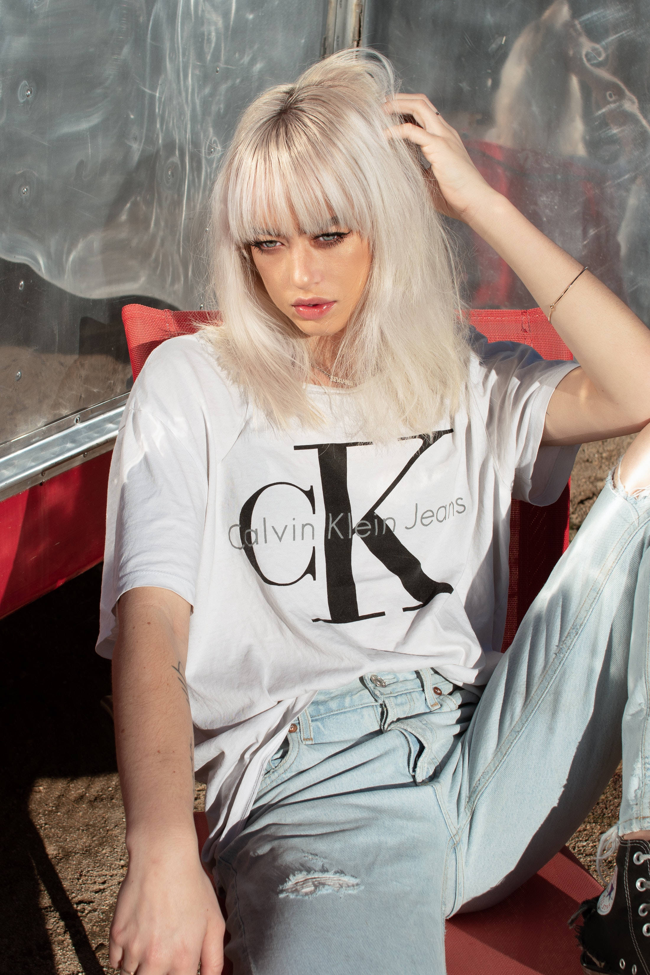 Klein 桌布 壁紙 — 一位年輕女性留著鉑金色頭髮，穿著Calvin Klein Jeans T恤和破洞牛仔褲，隨意地坐在紅色椅子上。