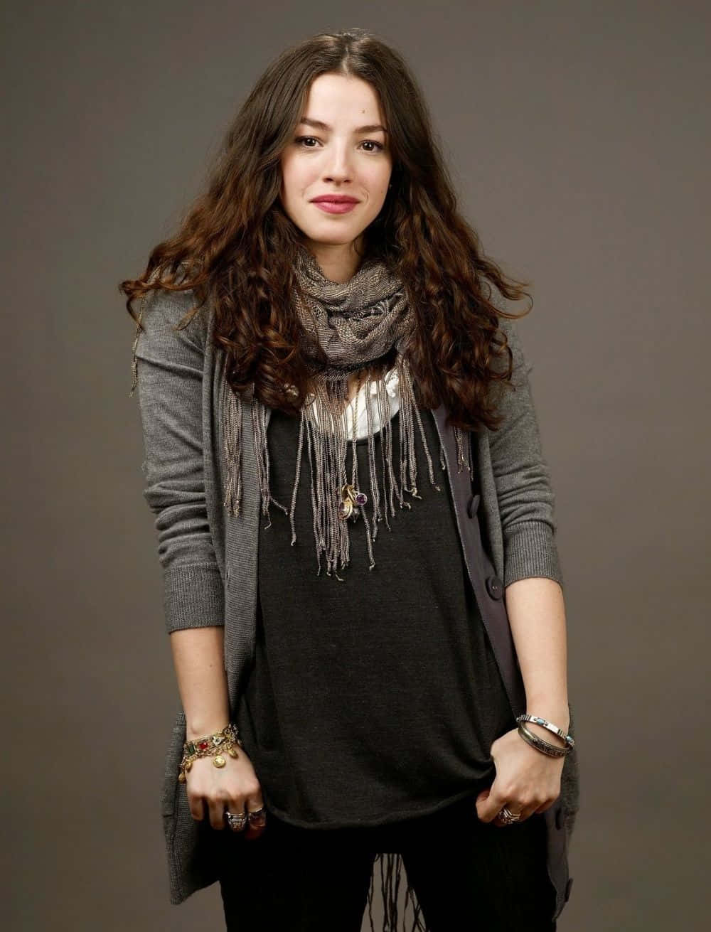 Olivia Thirlby 桌布 壁紙 — 一位年輕女性留著長長的波浪棕色頭髮，穿著灰色開襟衫和流蘇圍巾，背景是純灰色攝影棚。