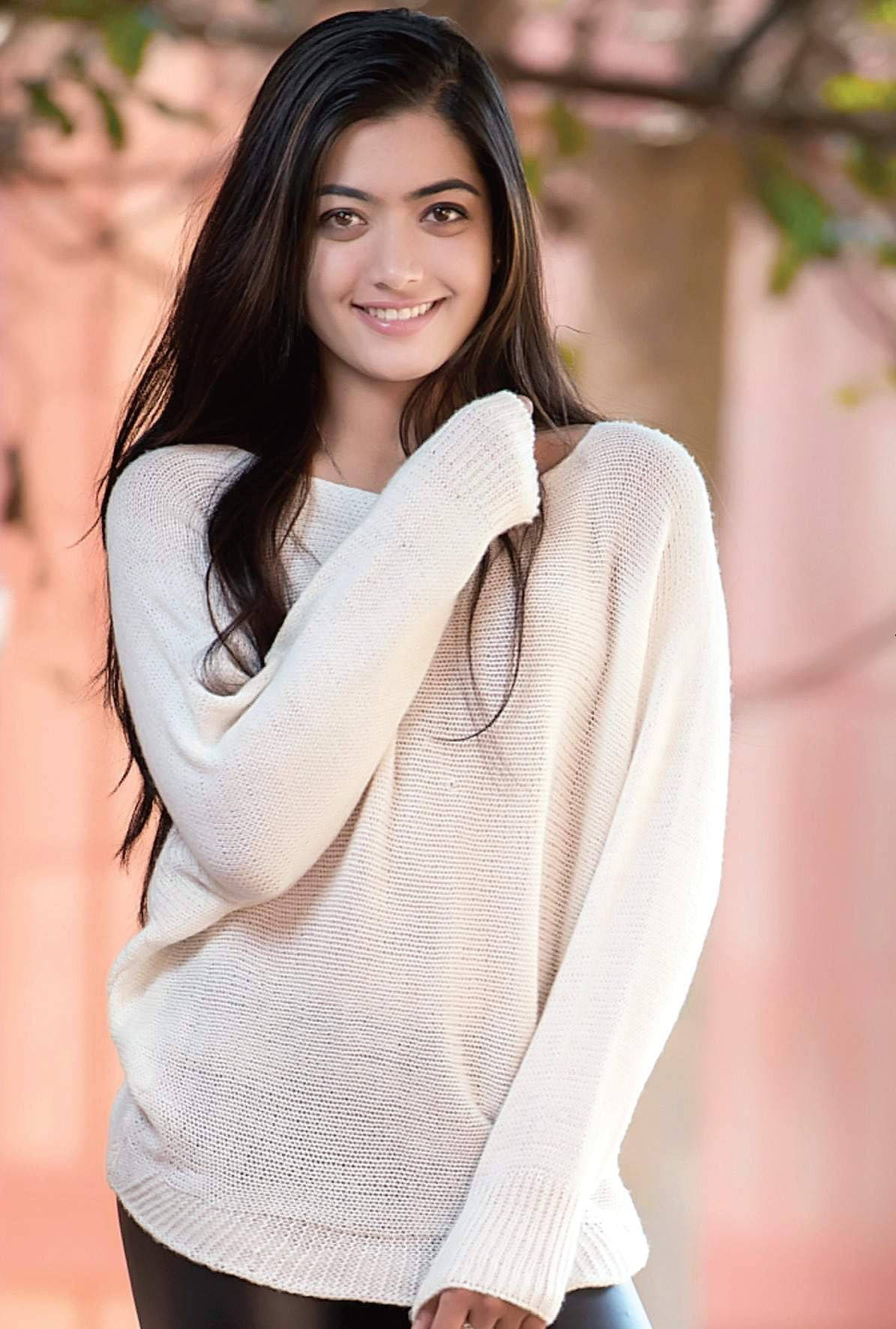 年輕女性穿著奶油色毛衣在戶外微笑，背景模糊。 — 拉什米卡·曼丹娜 壁紙