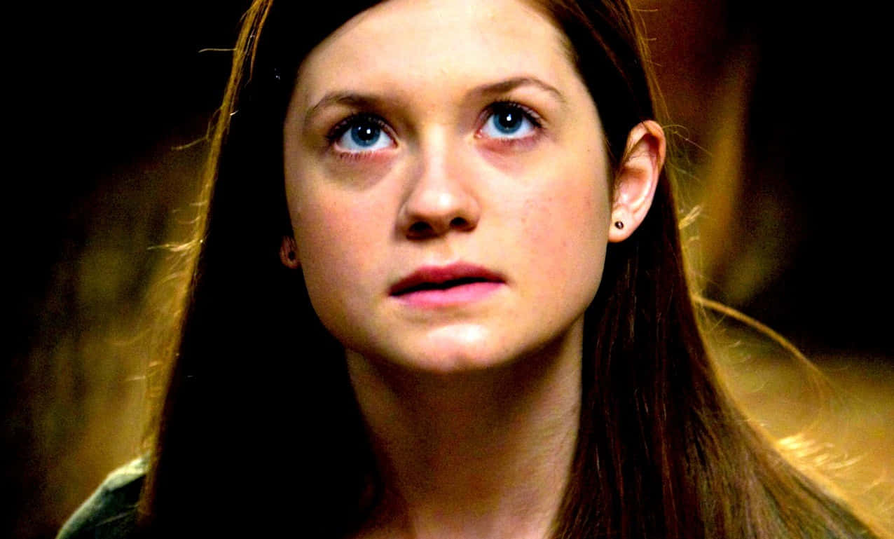 Ginny Weasley 桌布 壁紙 — 一位年輕女性的特寫肖像，擁有引人注目的藍色眼睛和長長的棕色頭髮，帶著嚴肅的表情向上凝視。