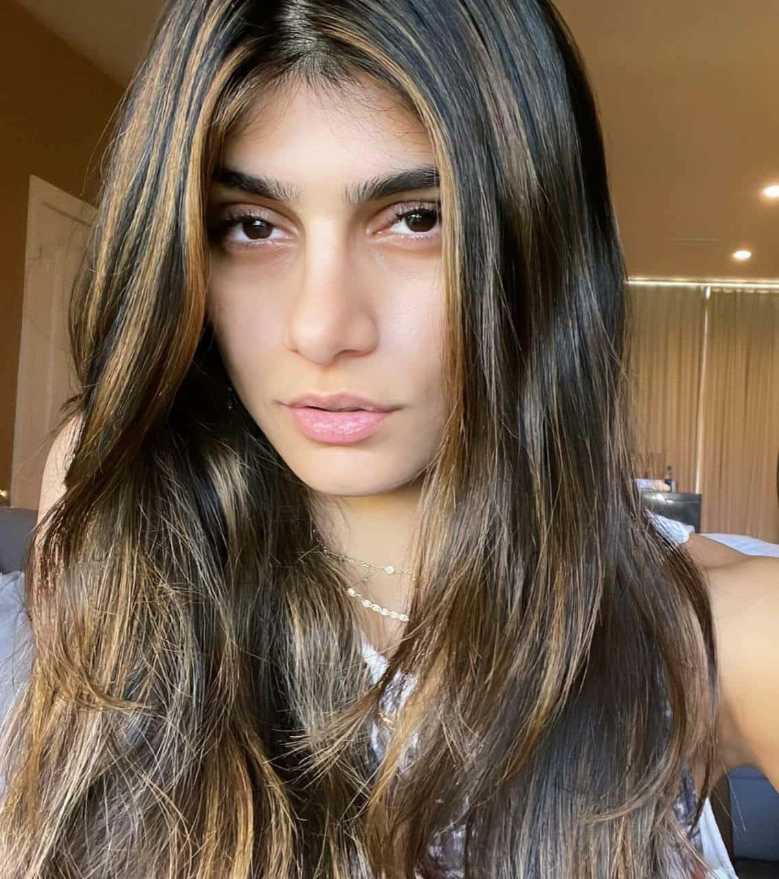 Mia Khalifa 桌布 壁紙 — 一位年輕女性的特寫肖像，擁有長而分層的棕色頭髮和柔和妝容，在室內直視鏡頭。