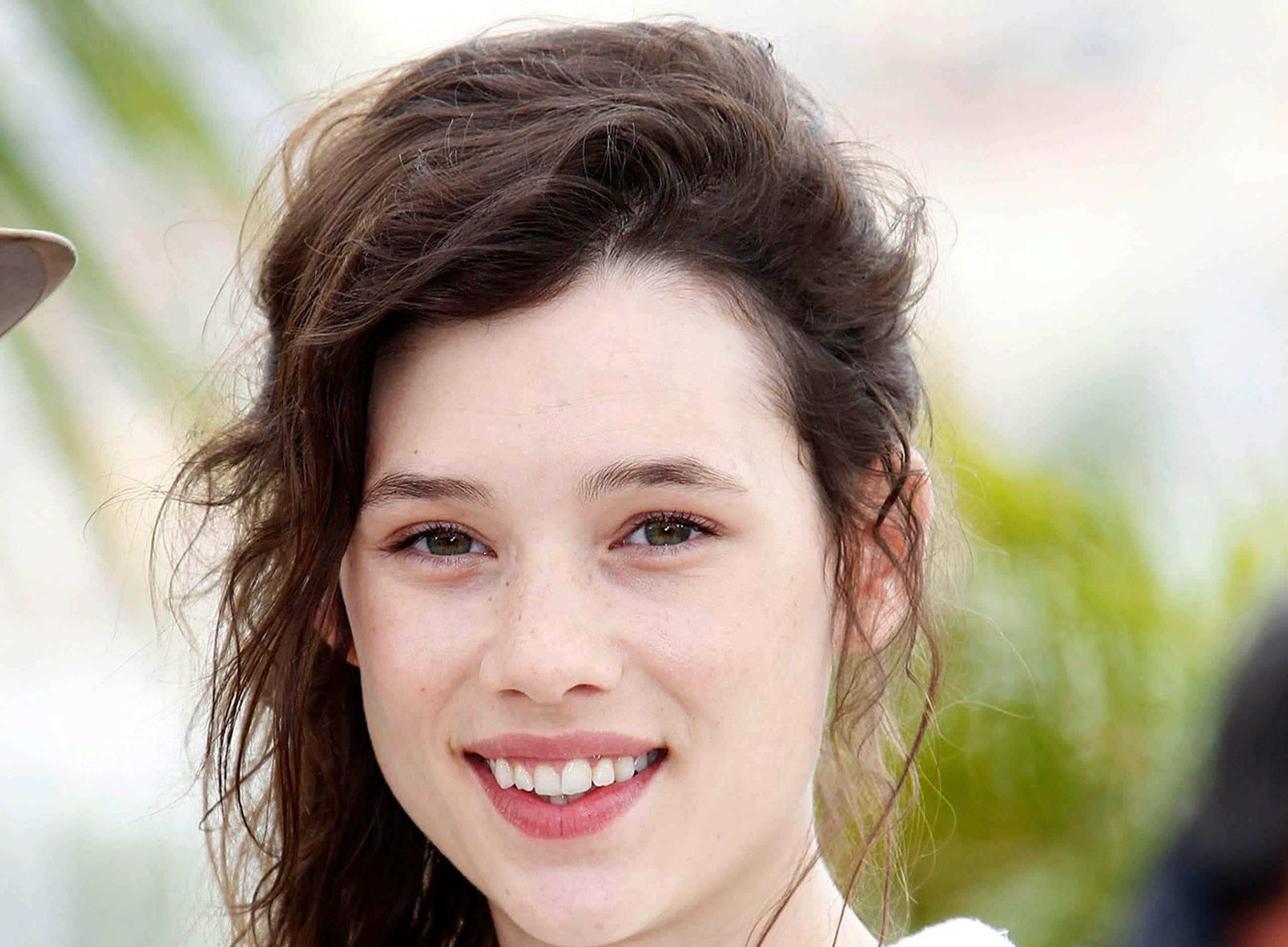 Astrid Berges Frisbey 桌布 壁紙 — 一位年輕女性的近距離肖像，她有一頭凌亂的棕色頭髮，在柔和模糊的綠色背景下溫暖地微笑。