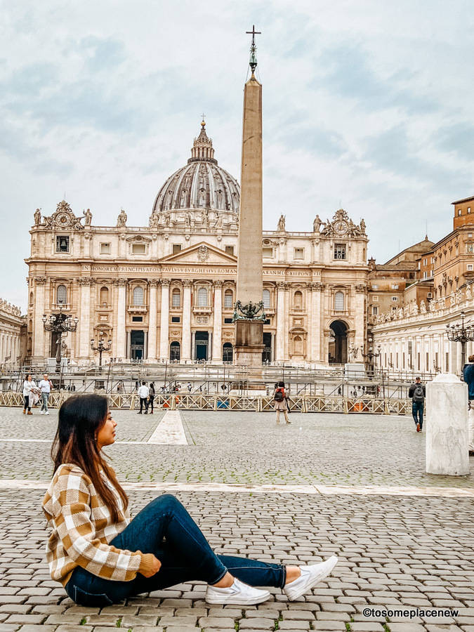 Rome 桌布 壁紙 — 一位年輕女性盤腿坐在鵝卵石上，凝視著陰天下的聖彼得大教堂。