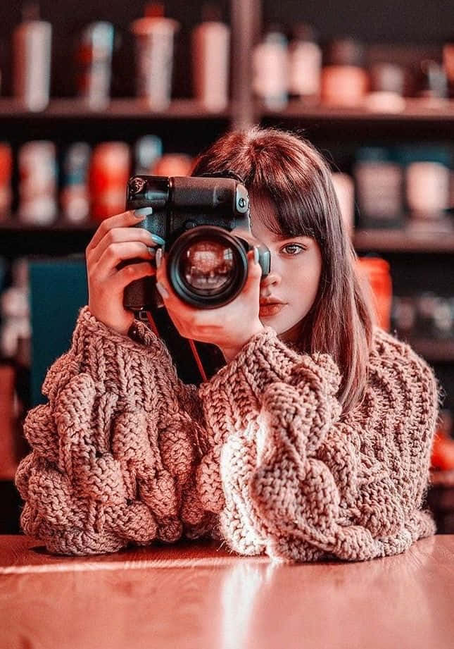 Holding Camera 桌布 壁紙 — 一位年輕女性穿著厚針織毛衣，坐在燈光溫暖的咖啡廳木桌旁，將相機舉到臉前。