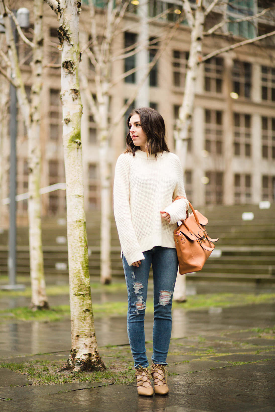 Minimalist Fashion 桌布 壁紙 — 一位年輕女性穿著奶油色毛衣和破洞牛仔褲，站在濕漉漉的城市公園中光禿禿的樹木旁，手持一個棕褐色背包。