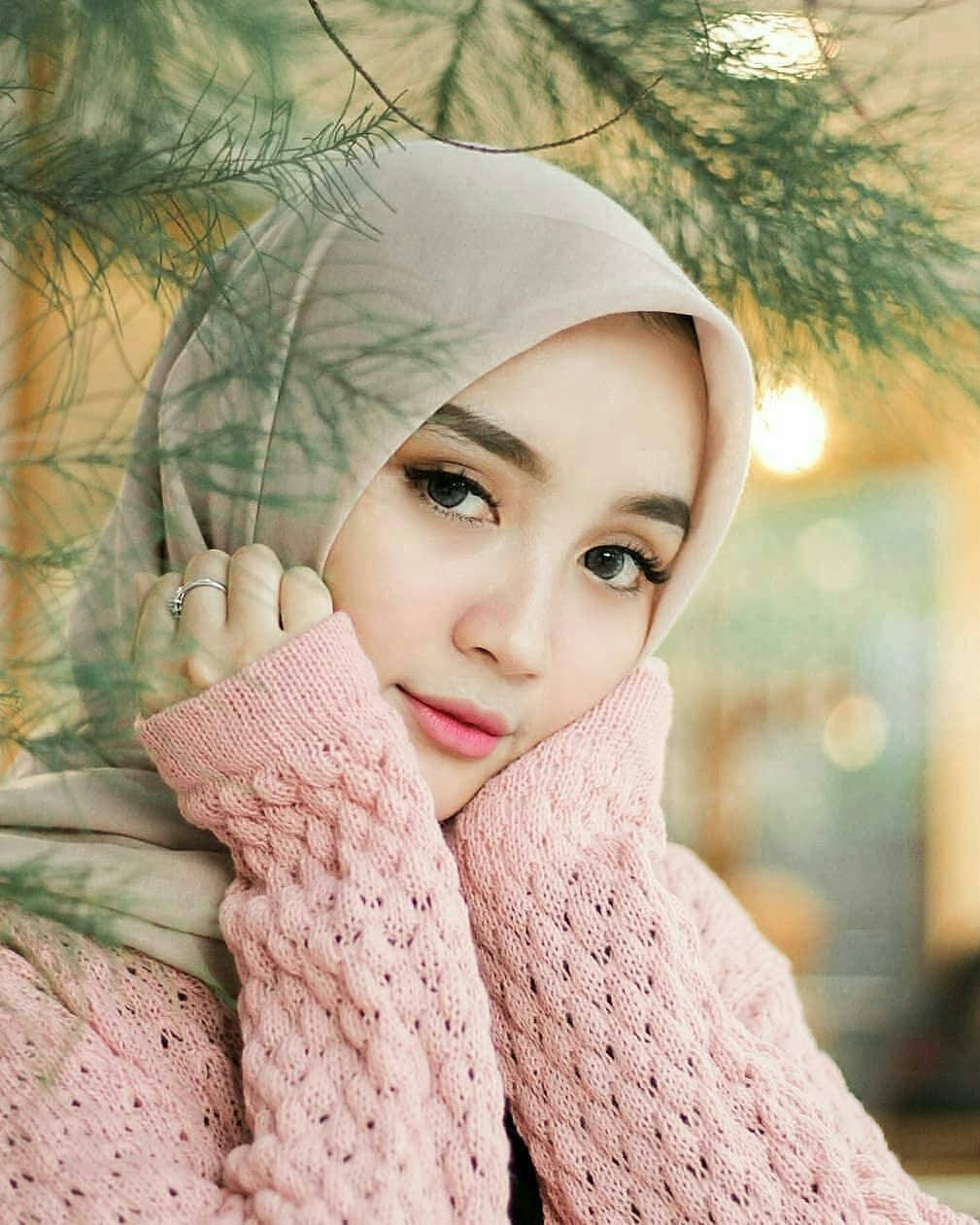 Cute Girl 桌布 壁紙 — 一位年輕女性穿著柔軟的米色頭巾和粉紅色針織毛衣，在綠色松枝下用手托著下巴。