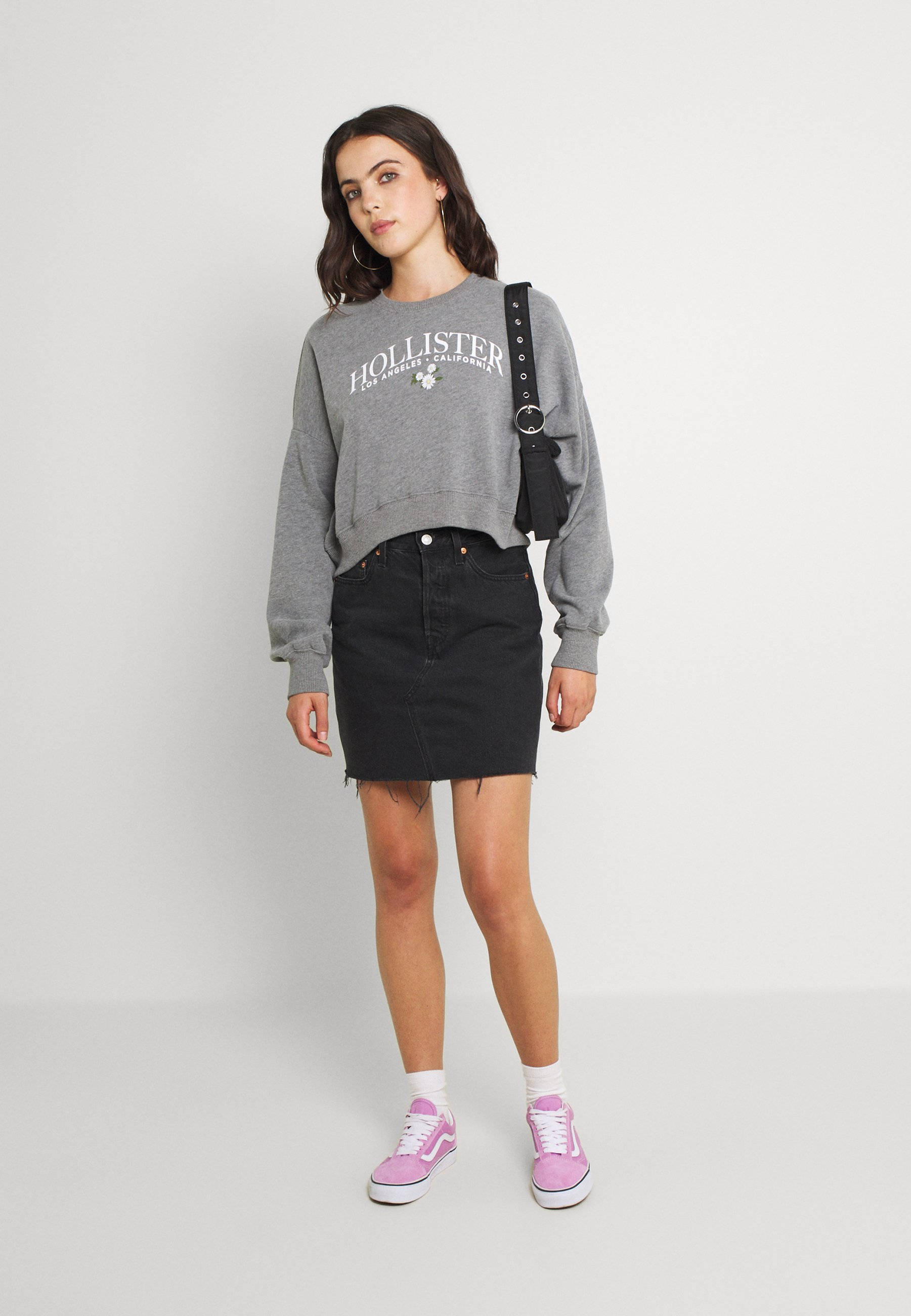 Hollister 桌布 壁紙 — 一位年輕女性穿著灰色Hollister連帽衫、黑色牛仔裙和粉紅色運動鞋，在純白色攝影棚背景前擺姿勢。