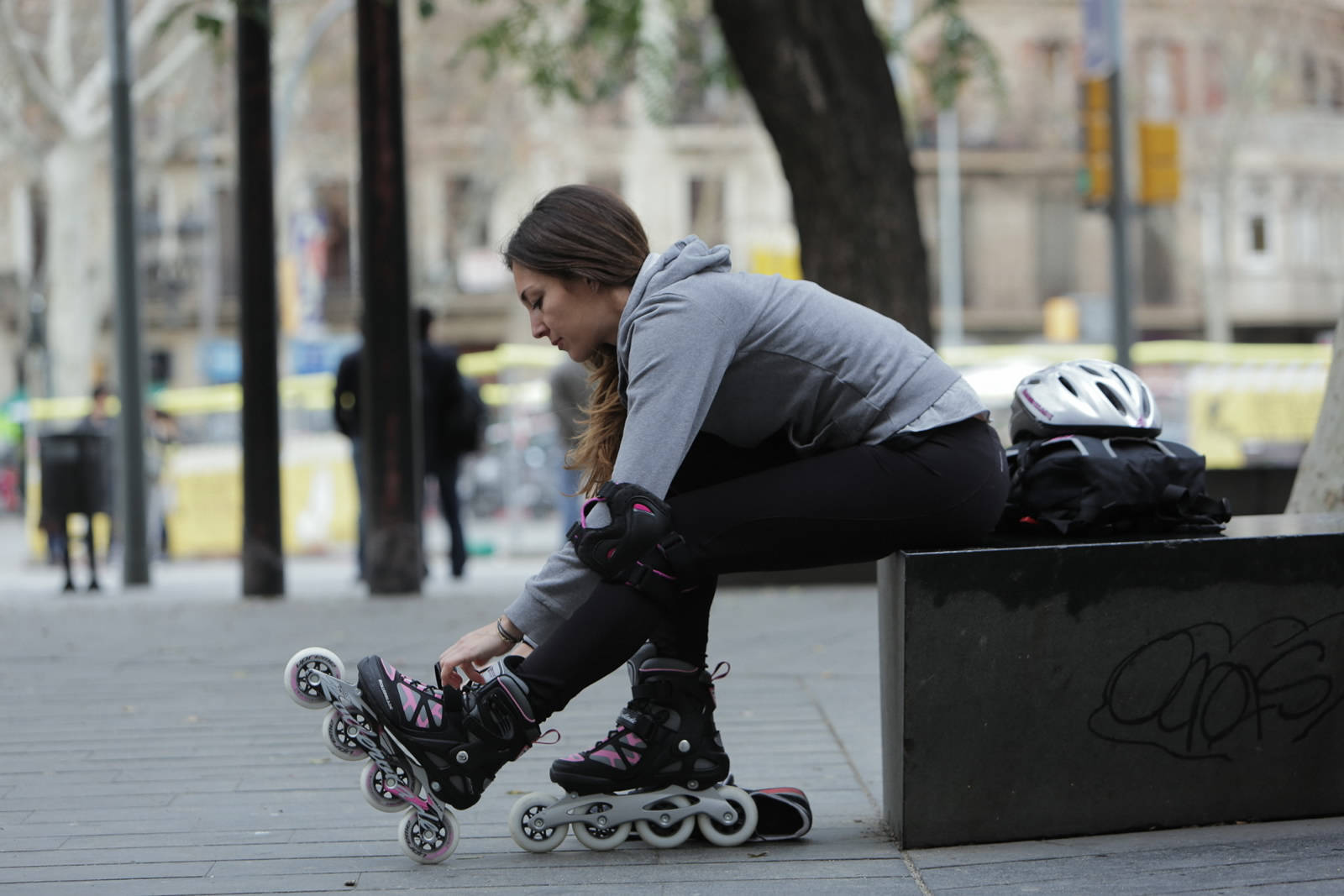 skating 桌布 壁紙 — 一位年輕女性穿著灰色連帽衫和黑色緊身褲，坐在城市廣場上有塗鴉的長椅上，調整她的粉紅色和黑色直排輪鞋。