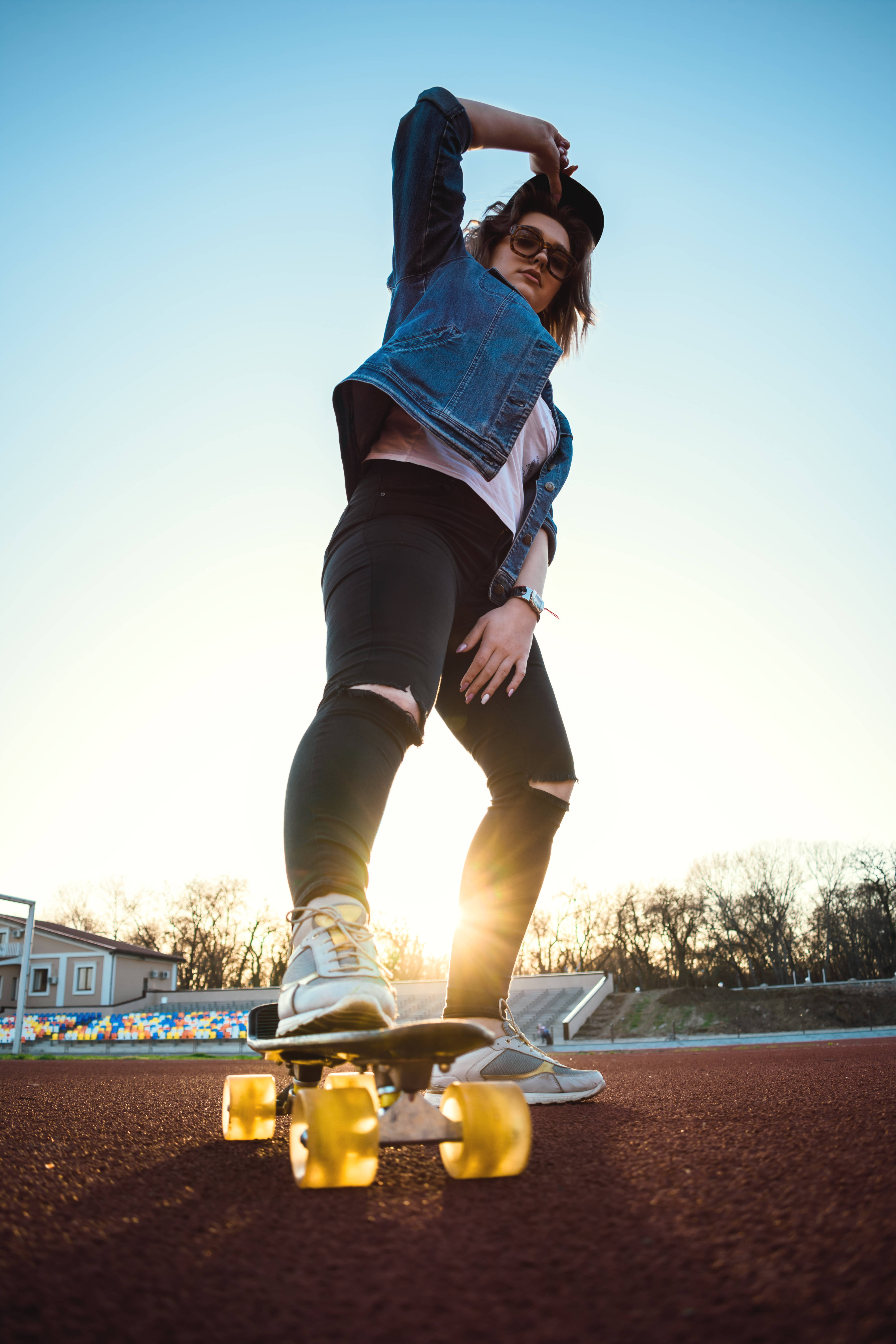Tomboy 桌布 壁紙 — 一位年輕女性穿著牛仔外套和太陽眼鏡，在日落時分於賽道上滑滑板，金色輪子發光。