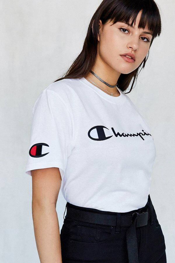 Champion Logo 桌布 壁紙 — 一位年輕女性穿著白色Champion T恤和黑色長褲，在純灰色背景前自信地擺姿勢。