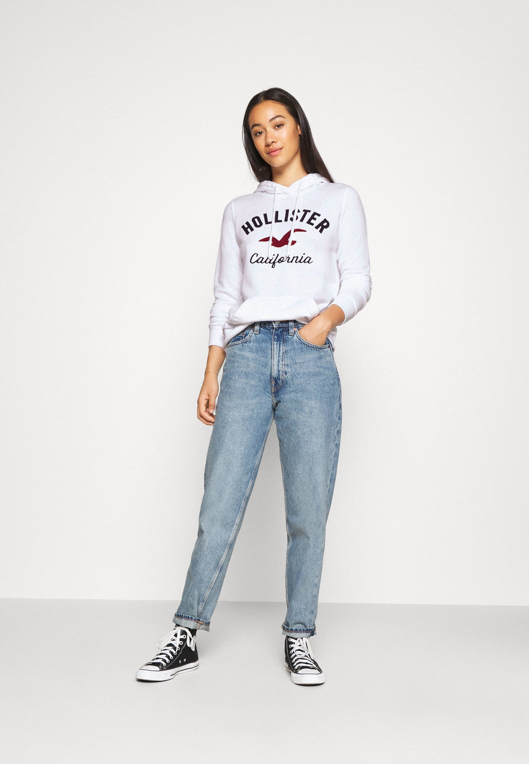 Hollister 桌布 壁紙 — 一位年輕女性穿著白色Hollister連帽衫、藍色牛仔褲和黑色運動鞋，在純白色攝影棚背景前自信地擺姿勢。